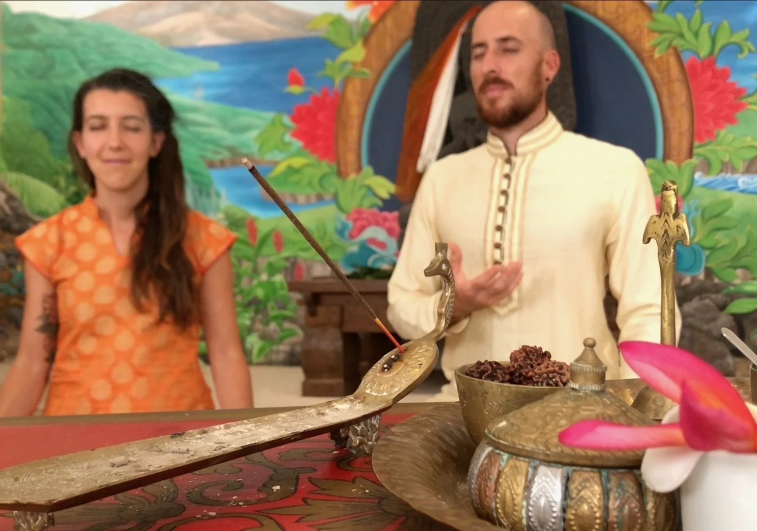 Two individuals participating in a spiritual or ritual ceremony, with one person holding a bow-shaped object and another with a hand placed on their chest, surrounded by colorful decorations and ritual items on a table.