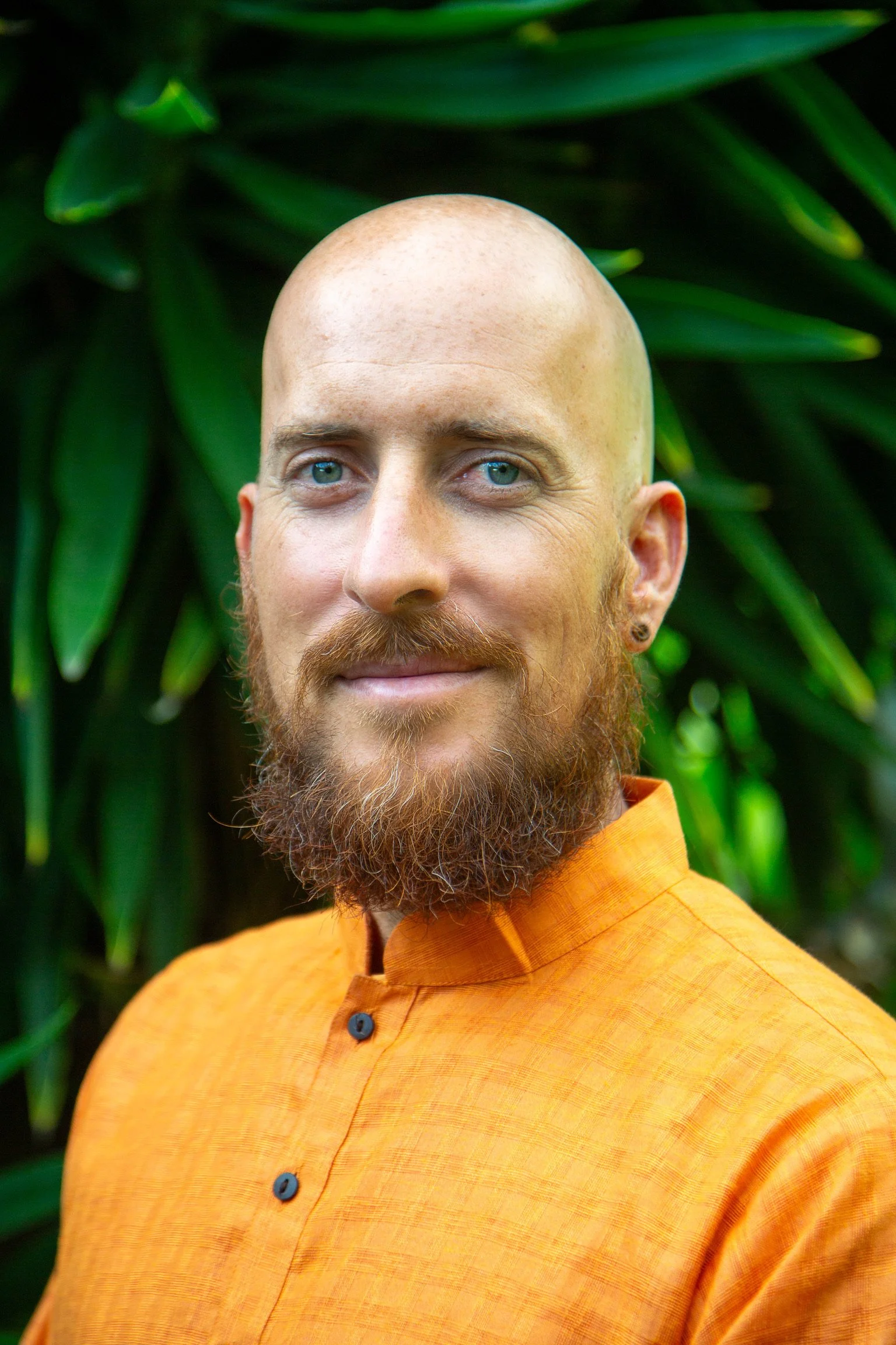 A male yoga instructor with a bald head and full beard wearing an orange shirt, standing in front of green leafy plants.
