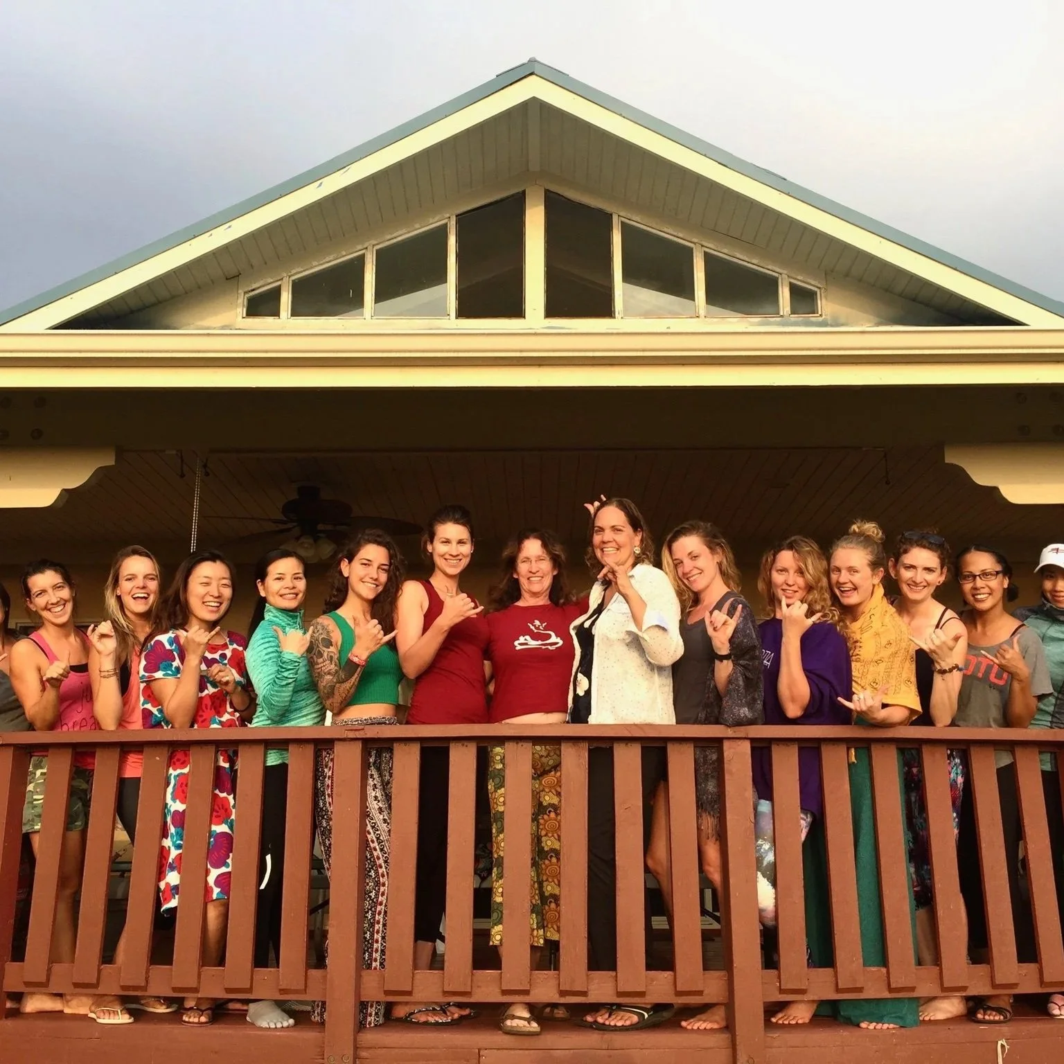 Group of women on a wooden balcony in front of a house, smiling and making hand gestures for friendship and fun.