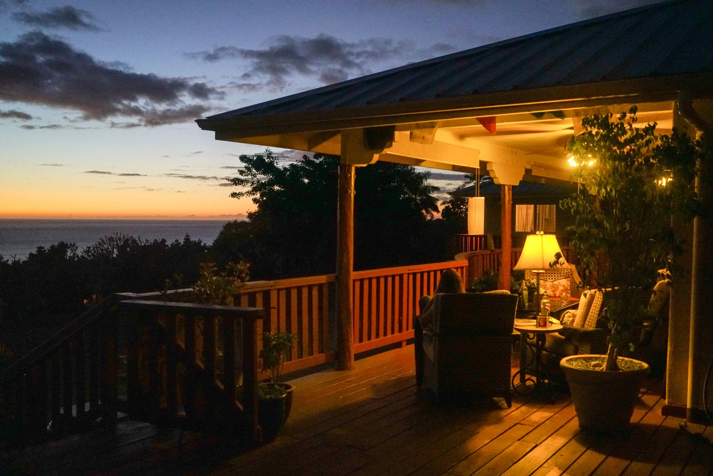 A warmly lit deck overlooks the ocean at dusk with tropical vegetation silhouettes in the background.