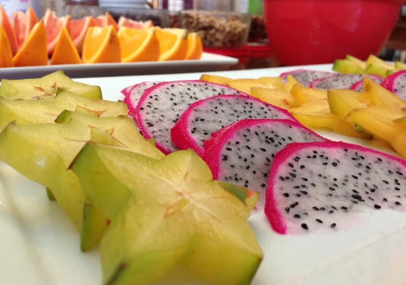 Close-up of sliced starfruit, dragon fruit, and other fresh fruits on a white platter with background of orange slices and other fruit