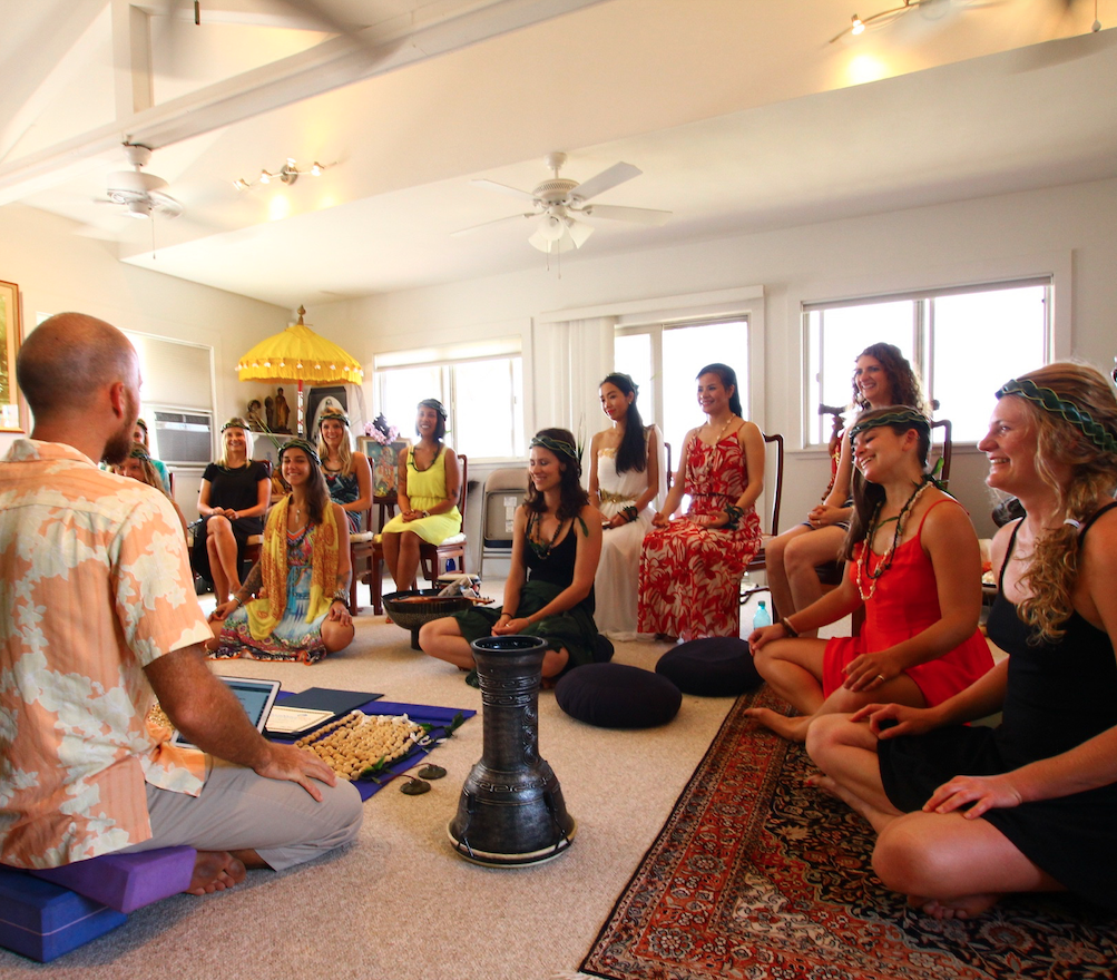 A line of yogis raise their arms to the sky while holding hands on a covered deck overlooking the ocean.