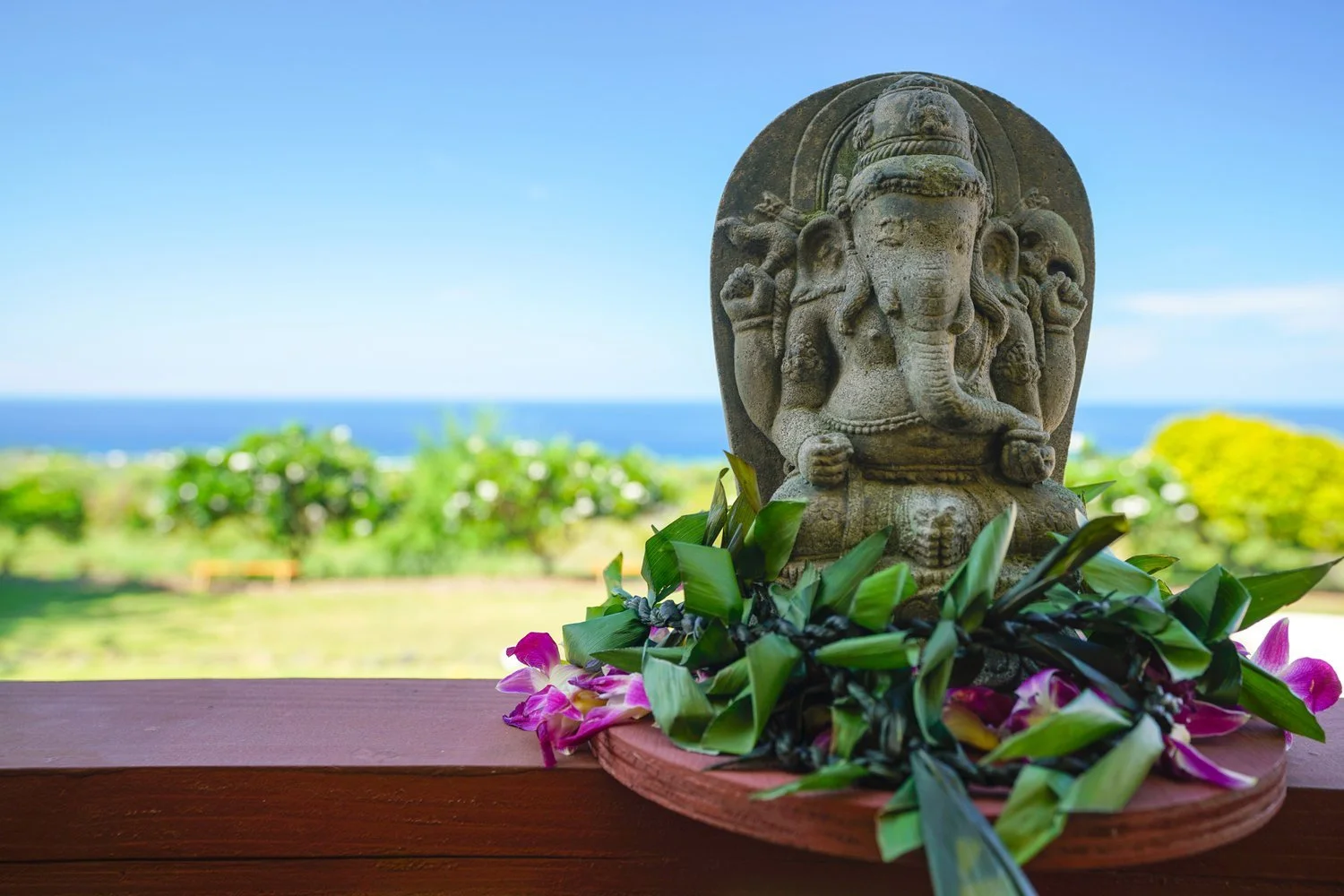 Stone sculpture of Ganesha with floral decorations, overlooking the ocean on a bright day.