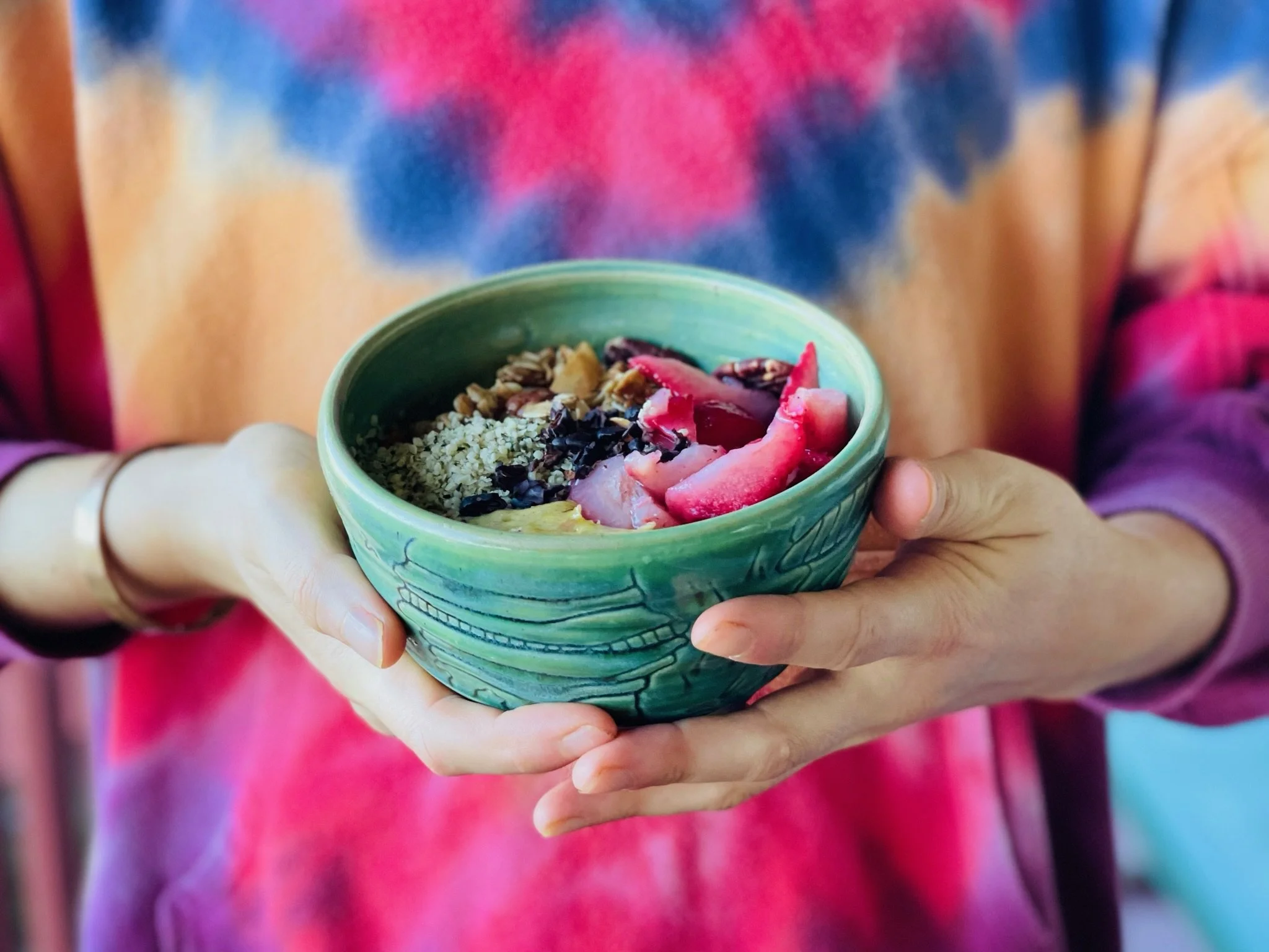 Close-up of a person holding a small, green, decorative bowl with a yogurt parfait topped with granola, fresh strawberries, blueberries, and possibly honey or syrup, with bright, colorful clothing in the background.