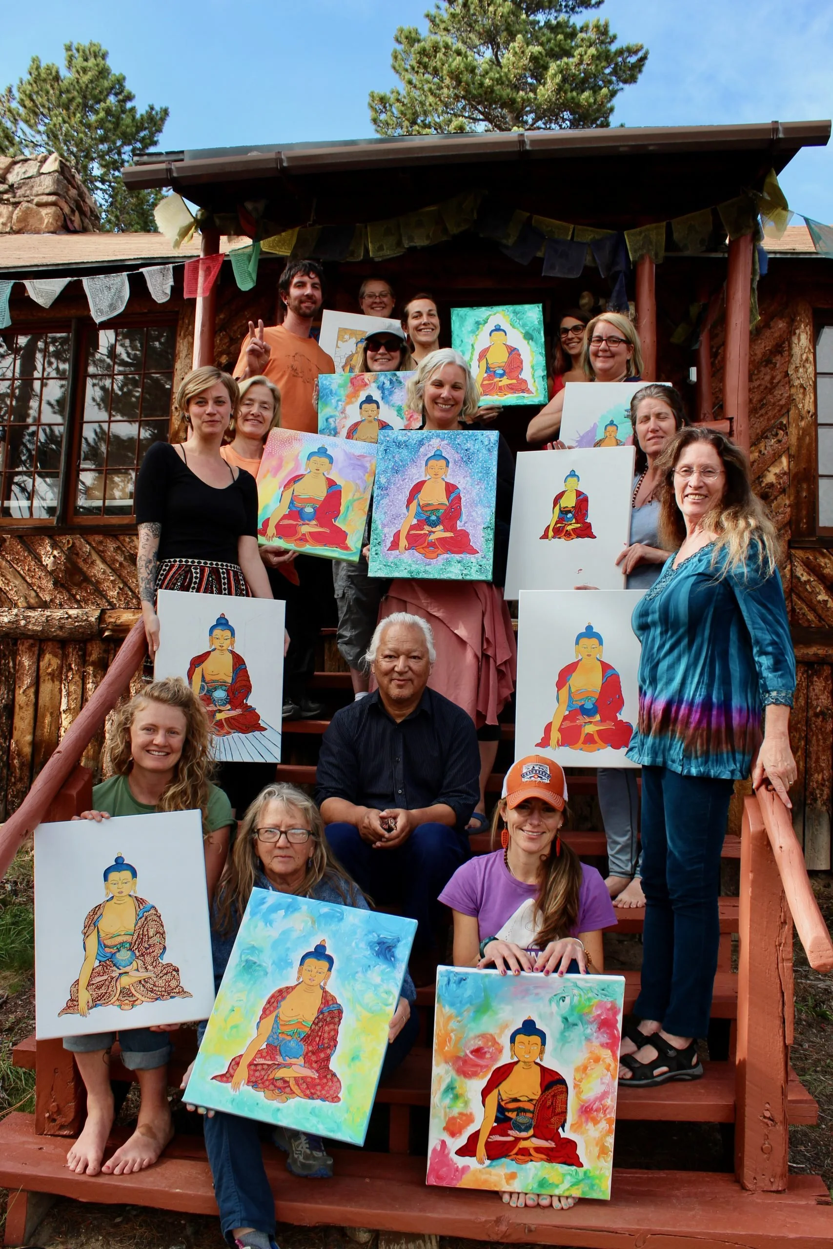 Group of people on outdoor stairs holding paintings of Buddha, with trees and a wooden cabin in the background.
