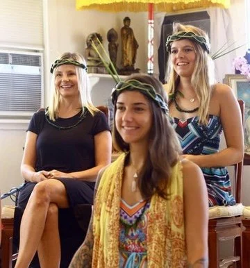 Three women wearing floral headbands seated and smiling during a gathering or celebration, with decorative items in the background.