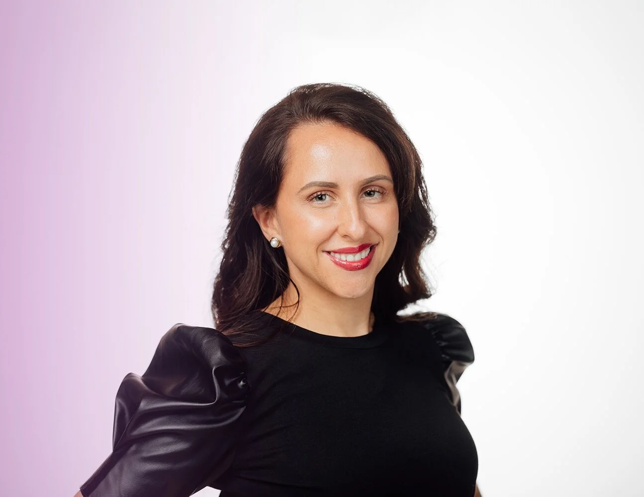 Smiling woman with long dark hair and pearl earrings wearing a black top with puffed sleeves, set against a white and purple gradient background.