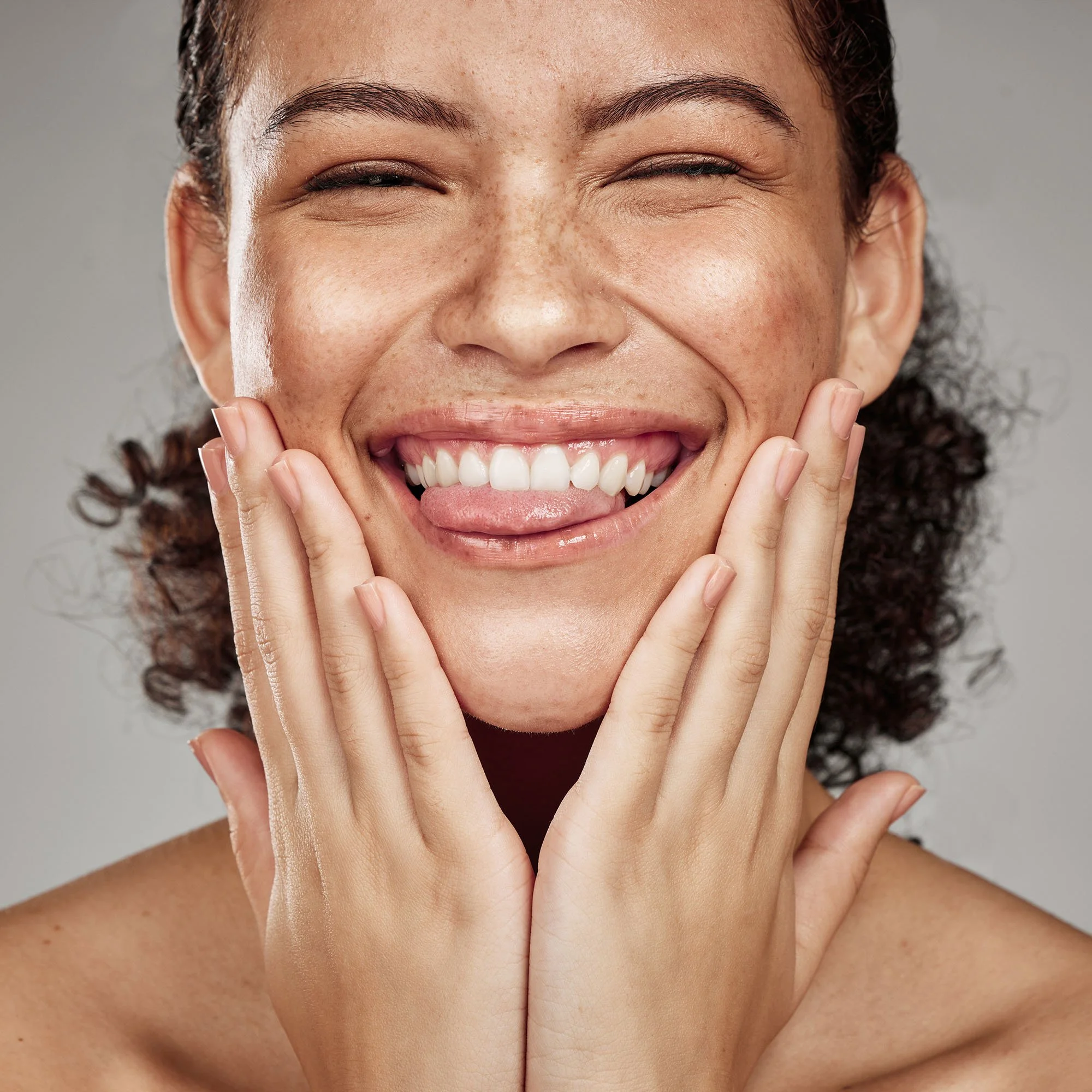 Close-up of a smiling woman with curly hair, touching her face with both hands, showing her teeth.