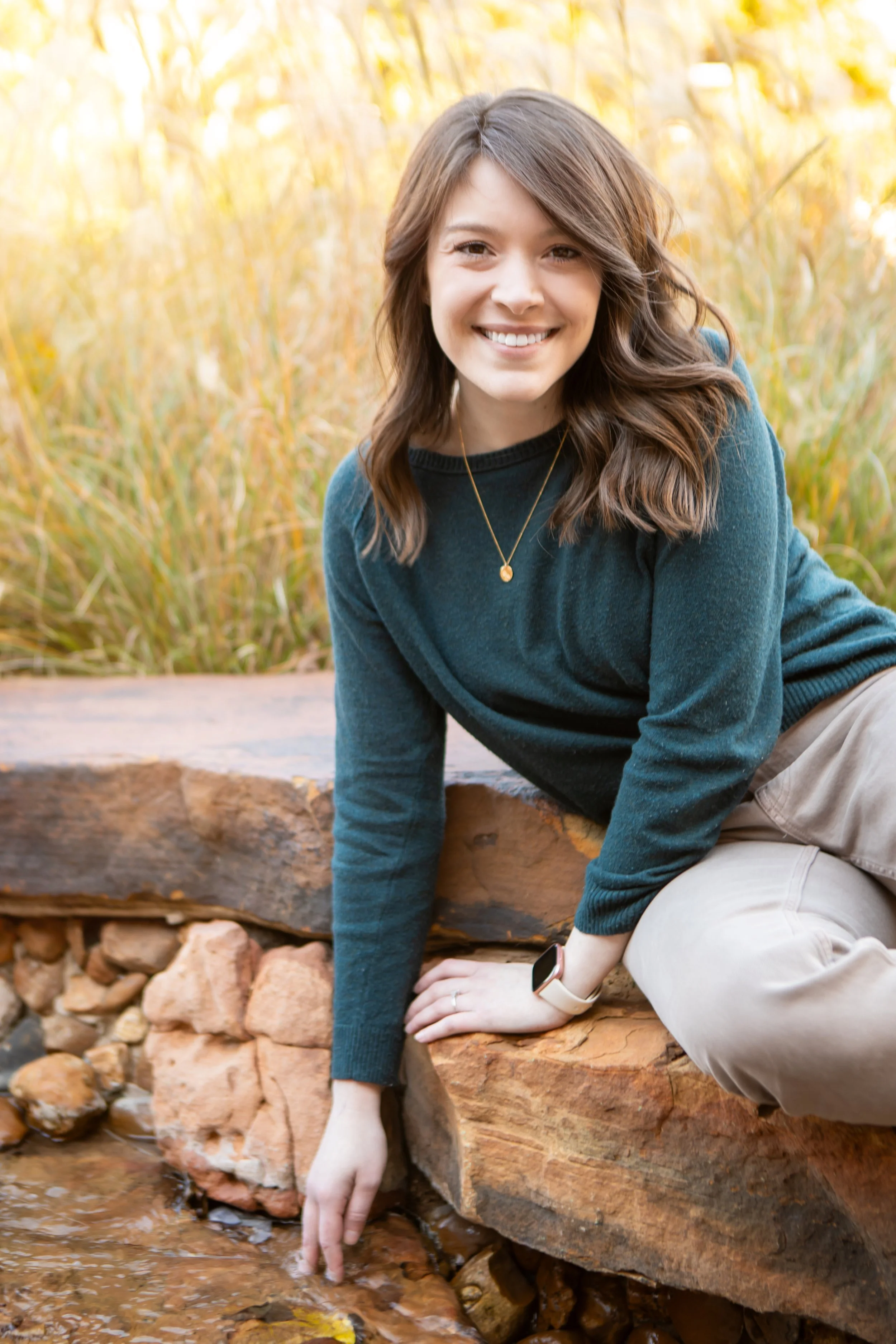 A woman whos is a licensed counselor with brown hair, wearing a teal sweater and beige pants, sitting on a rock near a small creek, touching the water with her hand. She is outdoors with tall yellowish grass in the background, during autumn.