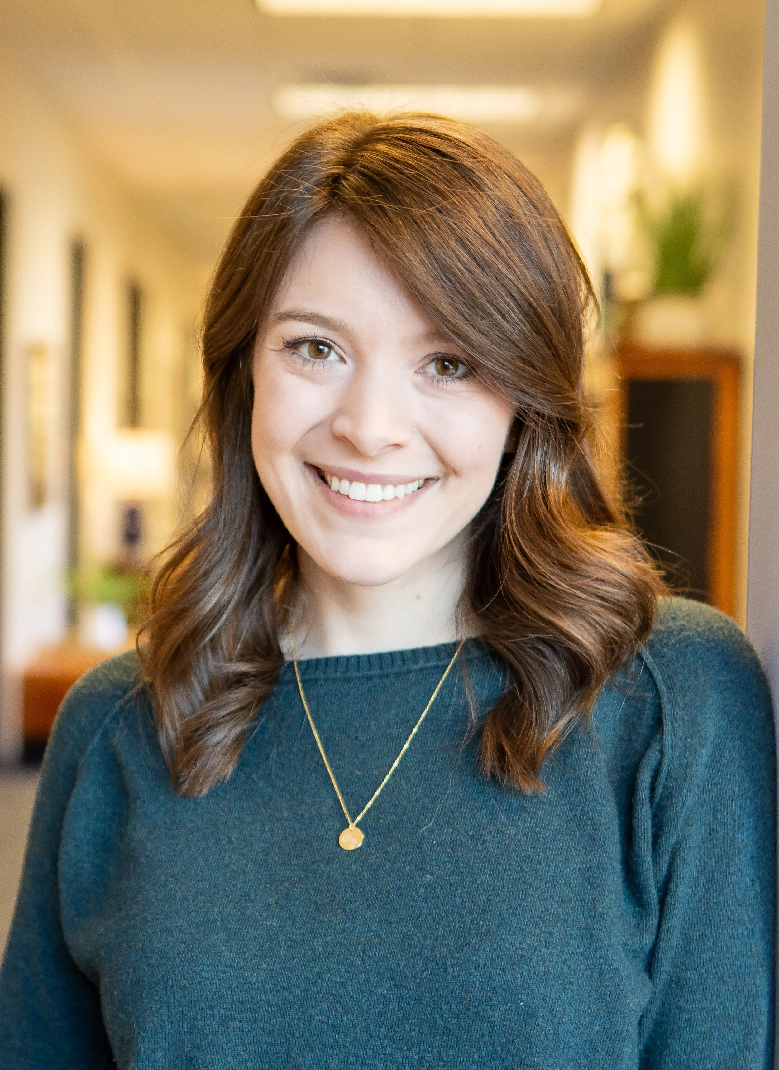 A young woman with shoulder-length brown hair and a bright smile, wearing a dark teal top and a gold necklace, standing in an indoor setting with framed pictures and plants in the background.