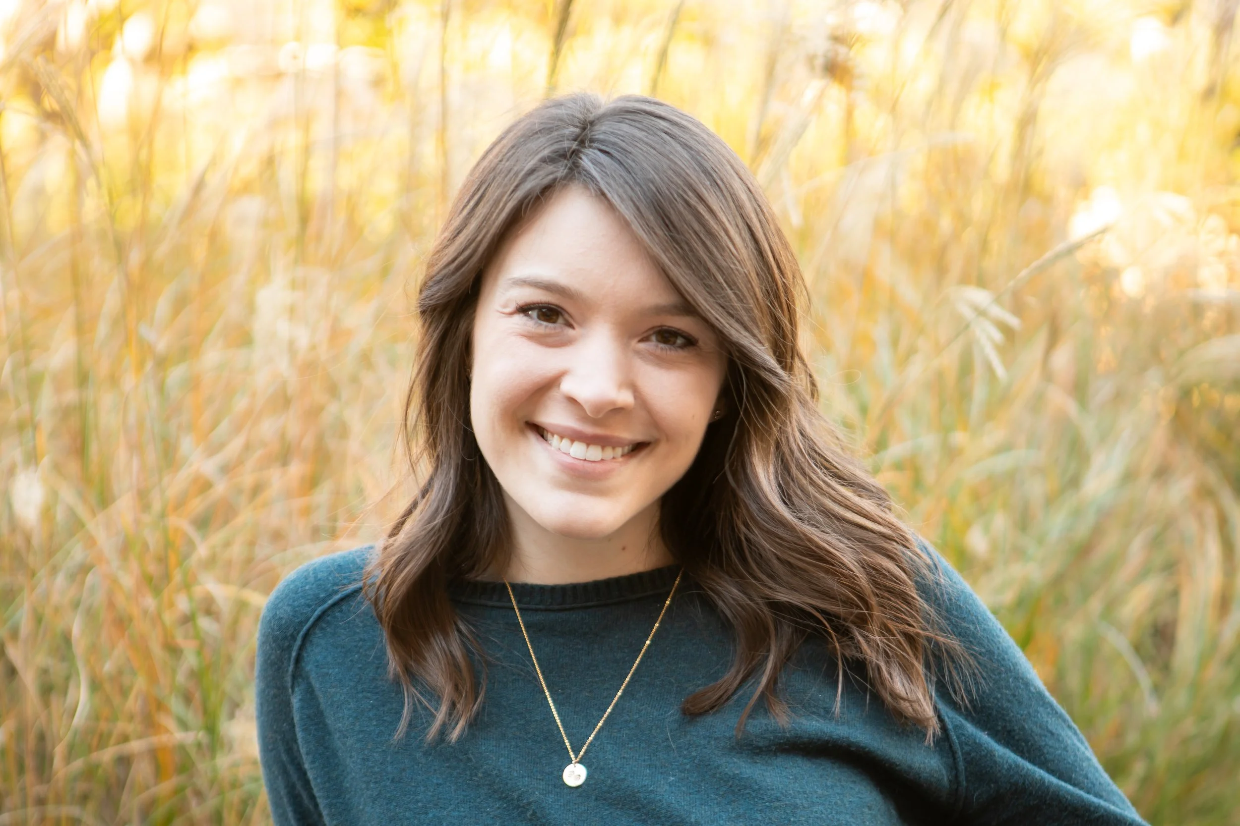 A young woman with brown hair smiling and wearing a blue sweater and a necklace, standing outdoors in a field of tall, golden grasses.
