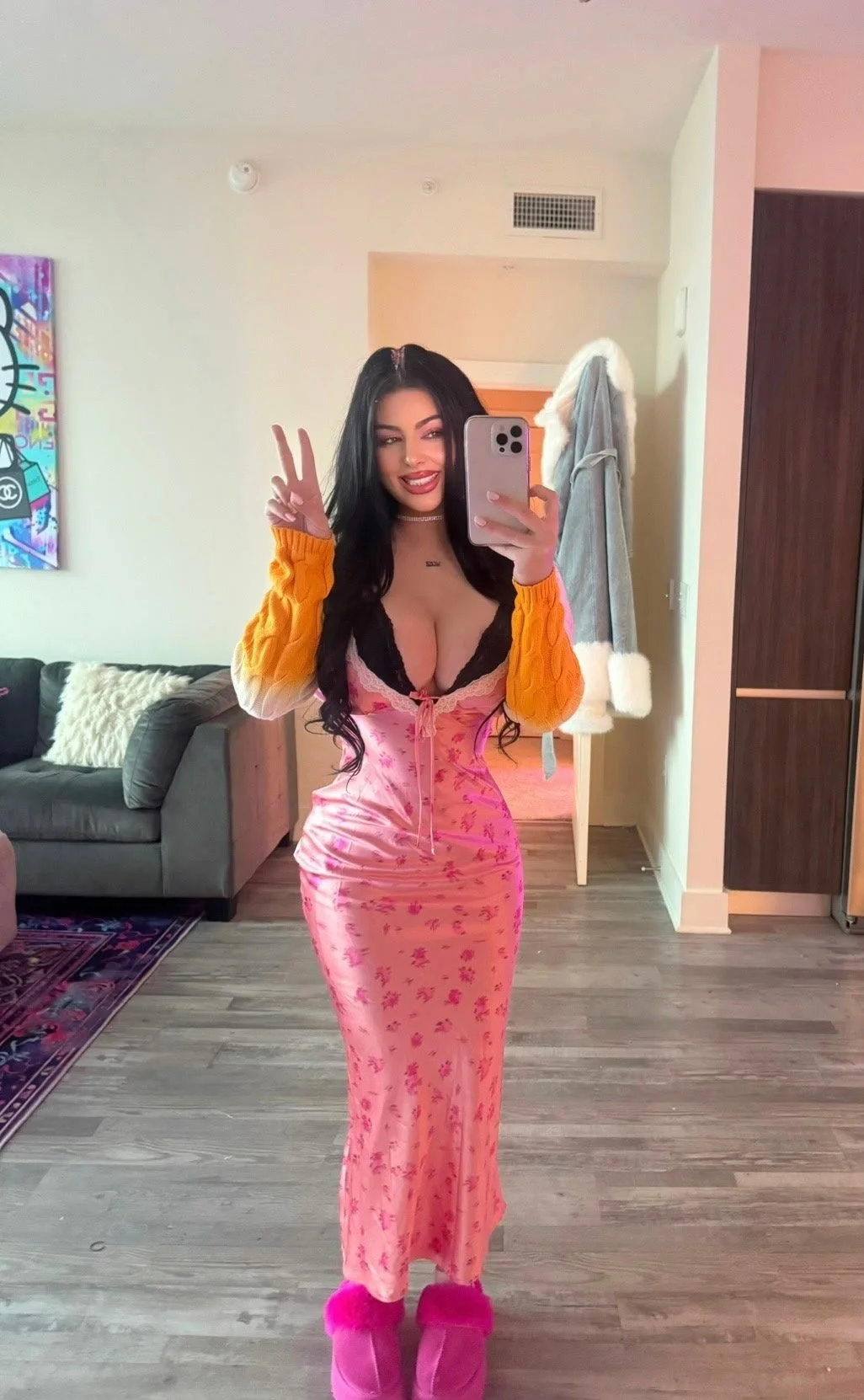 Person taking a mirror selfie in a room, wearing a floral dress, bright yellow sweater, and pink slippers, making a peace sign.