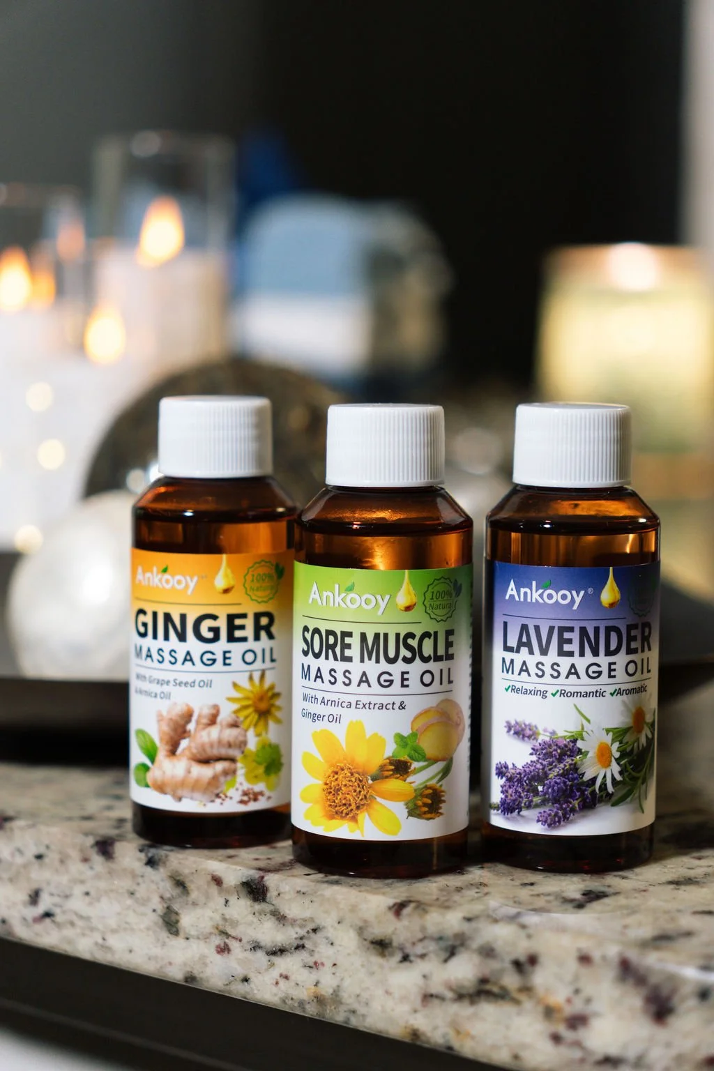Three bottles of Ankooy massage oil on a granite countertop; labeled Ginger Massage Oil, Sore Muscle Massage Oil, and Lavender Massage Oil, with relaxing background candles.