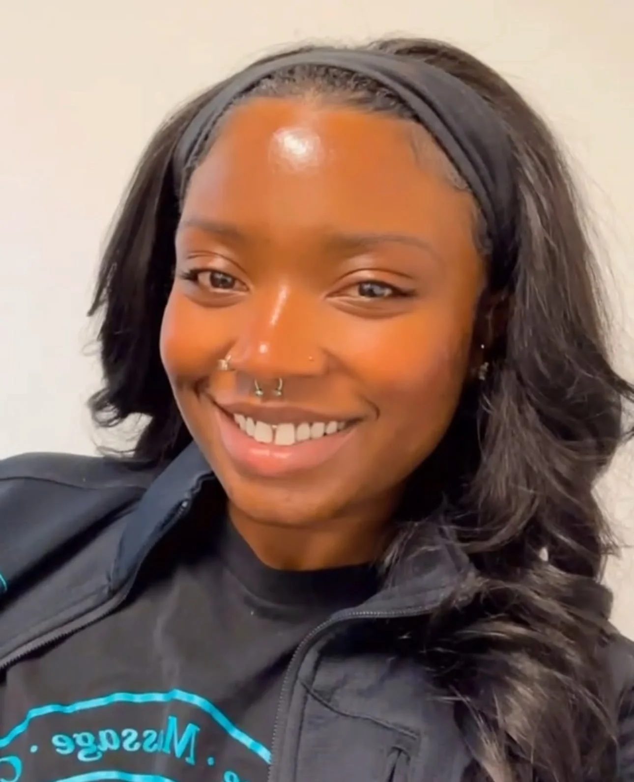 Smiling person with long hair, wearing a black headband, nose piercings, and a black jacket over a black shirt with blue text.