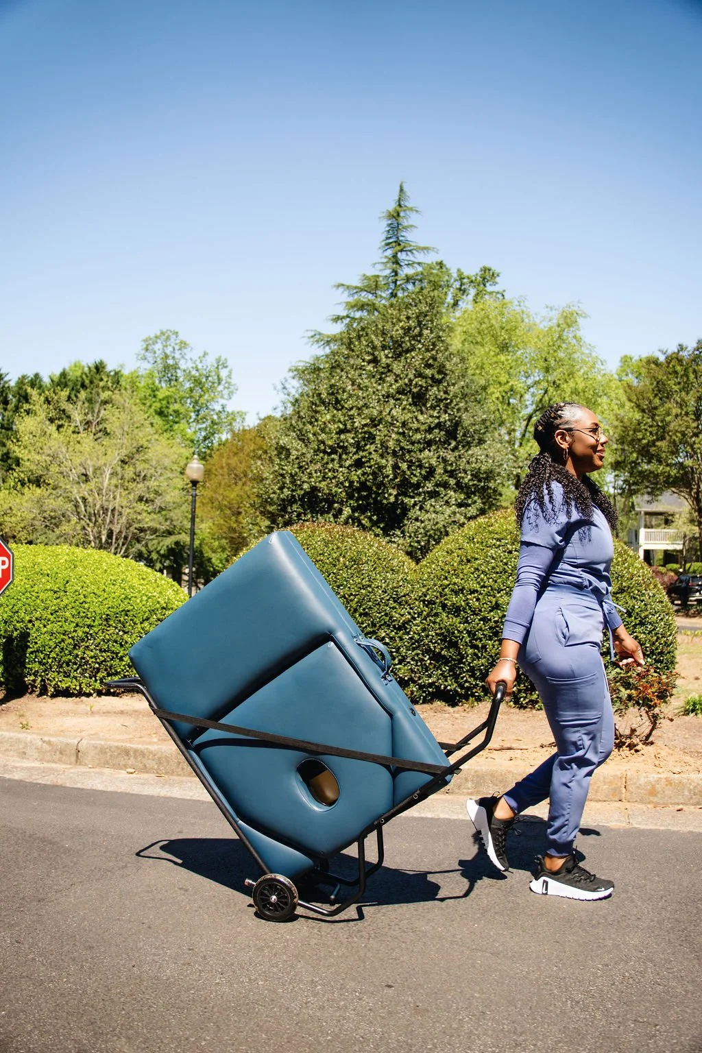 Person in blue massage therapy wear pulling a teal padded item on a wheeled cart outdoors.