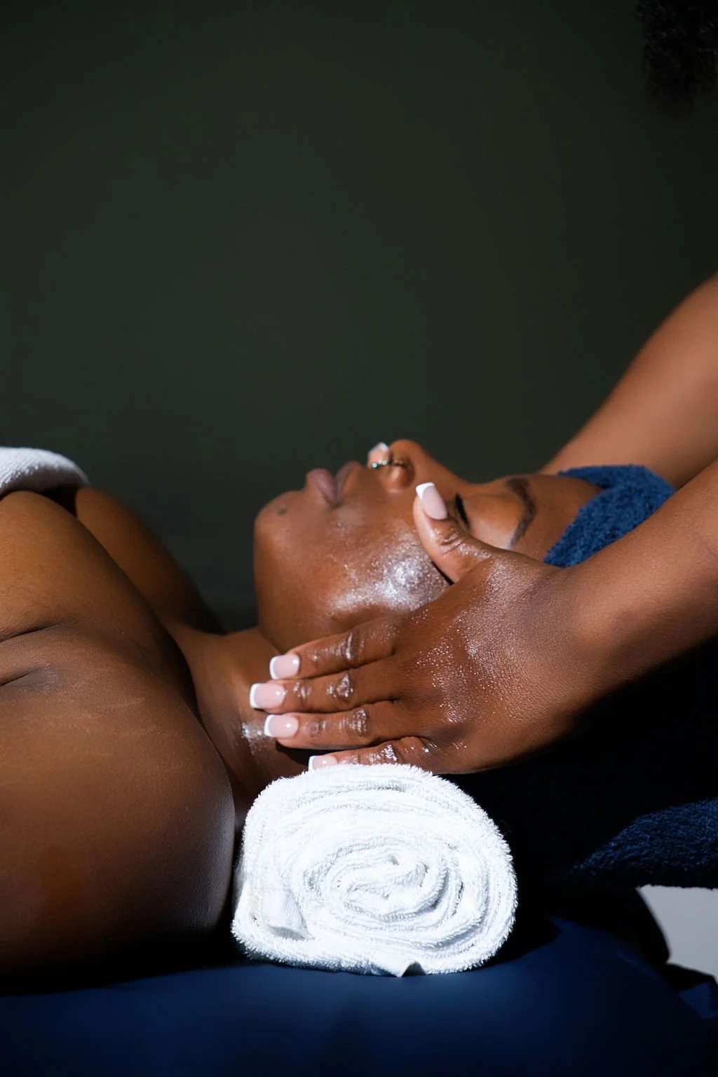 Person receiving a facial massage with hands applying oil, lying on a massage table with a rolled towel under their head.