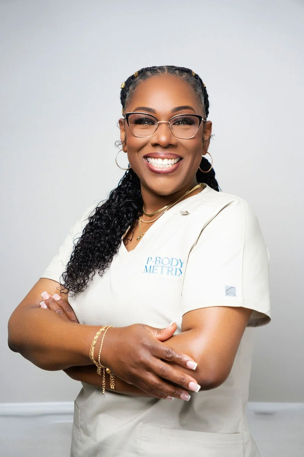 A woman smiling, wearing glasses and a uniform with the logo "P-Body Metrix." She has braided hair and is crossing her arms, with gold bracelets on her wrist. The background is plain and light-colored.
