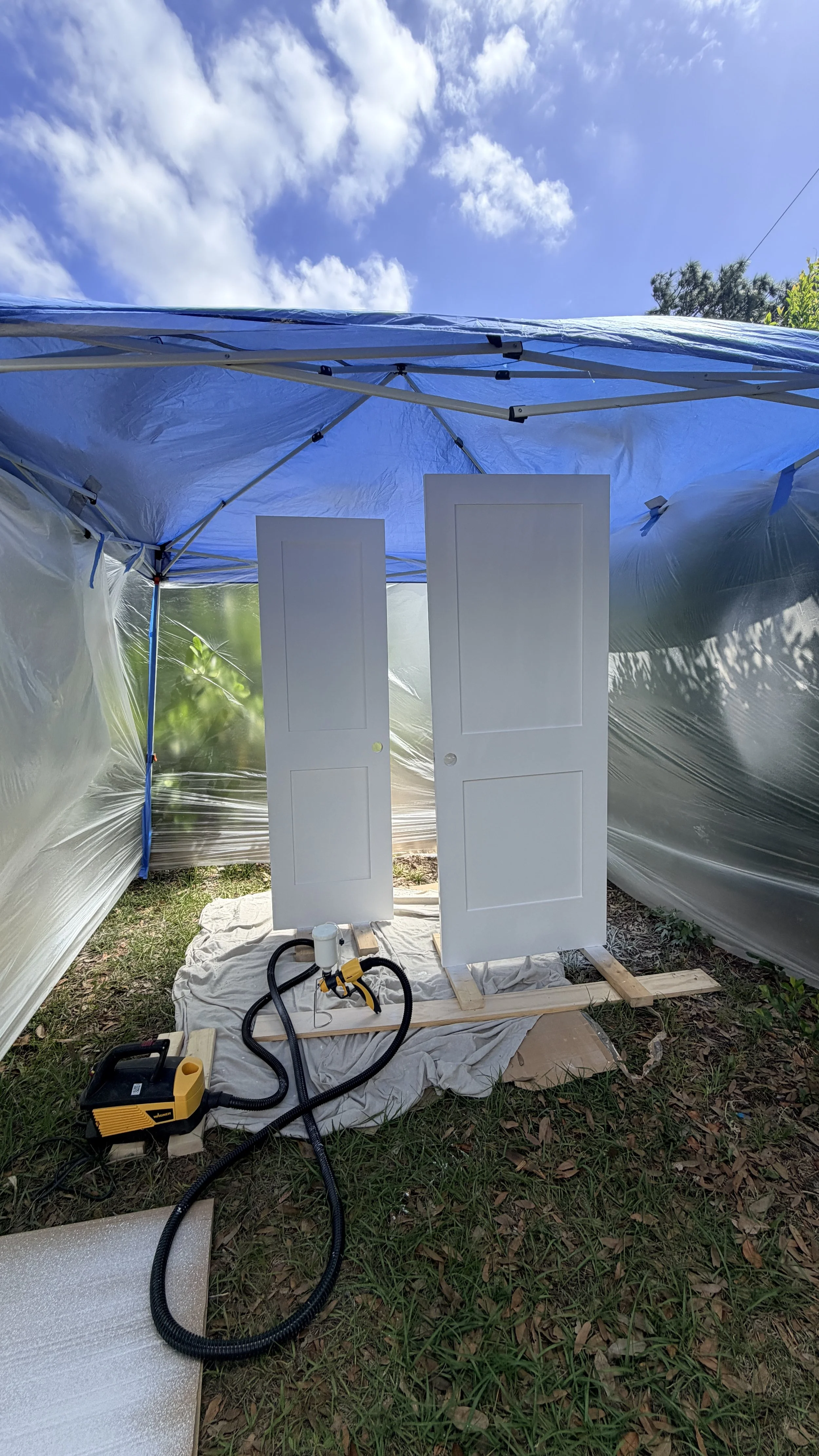 A partially assembled outdoor tent with a blue canopy, two white doors leaning against each other, and various tools and equipment on the ground, including a yellow heat gun and vacuum cleaner, set on a grassy area under a blue sky with clouds.