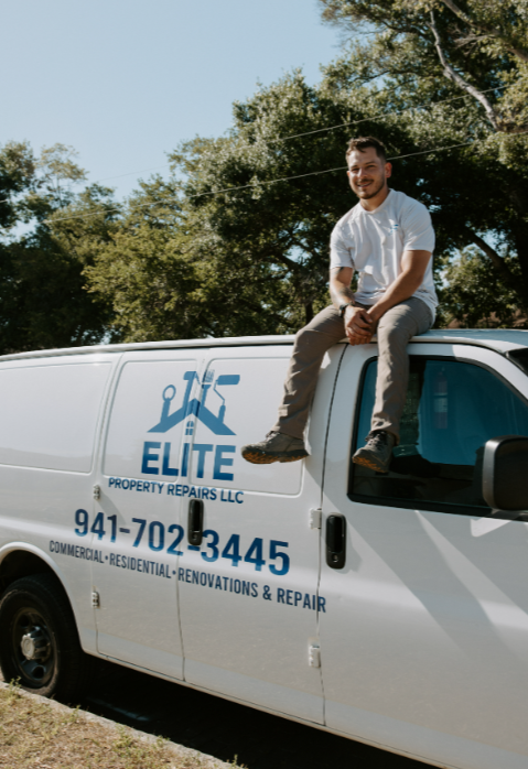 A man sitting on the hood of a white repair van outdoors with trees in the background, smiling at the camera.