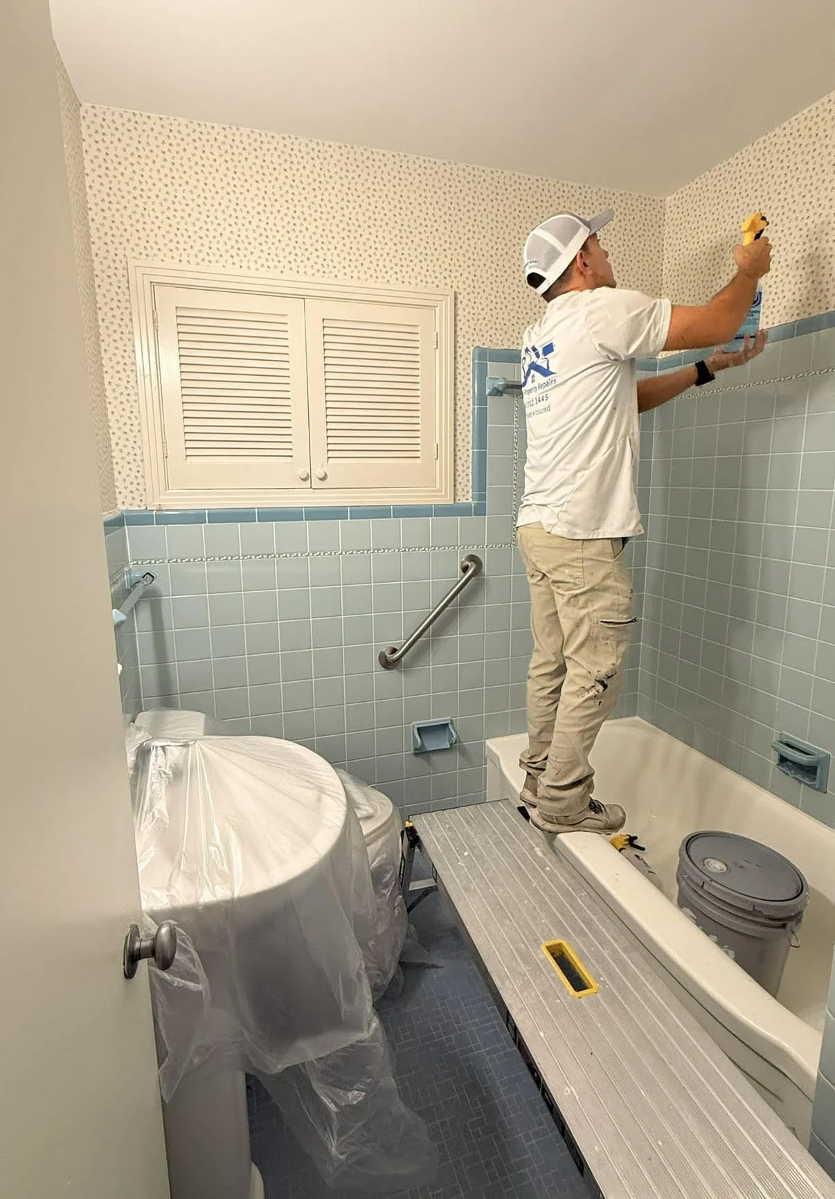 A man inspecting the upper corner of a bathroom wall using a yellow level and a blue spray bottle, surrounded by construction tools and covered fixtures.