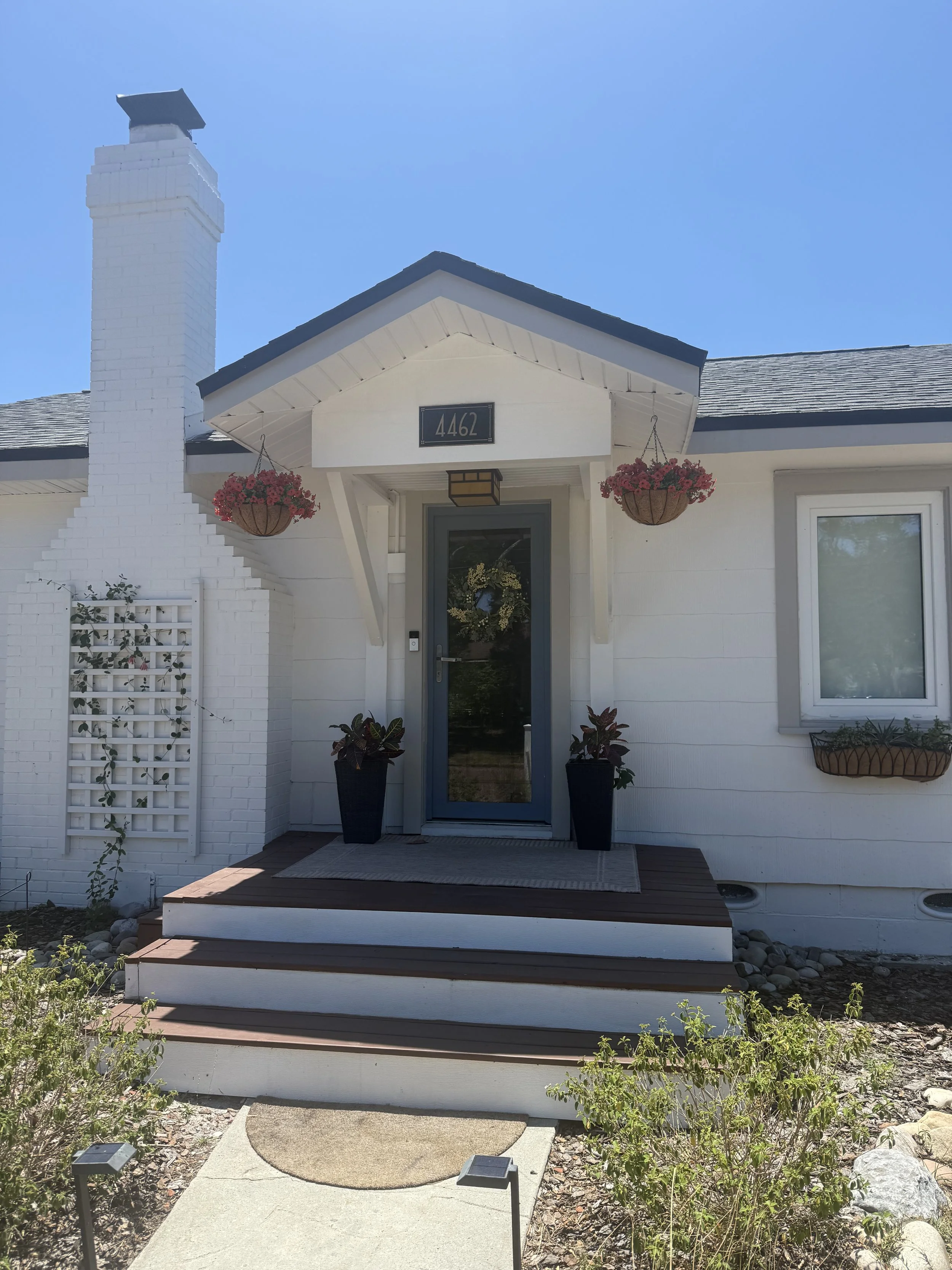 Front view of a white house with a gray front door, potted plants on either side, hanging flower baskets, and a small set of steps leading up to the entrance. The house has a black sign with the number 4462 above the door, and a chimney on the left s
