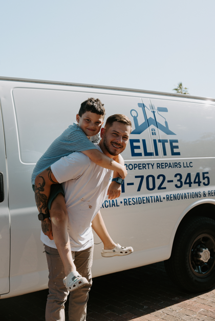 A man giving a piggyback ride to a young boy in front of a property repair company's van.