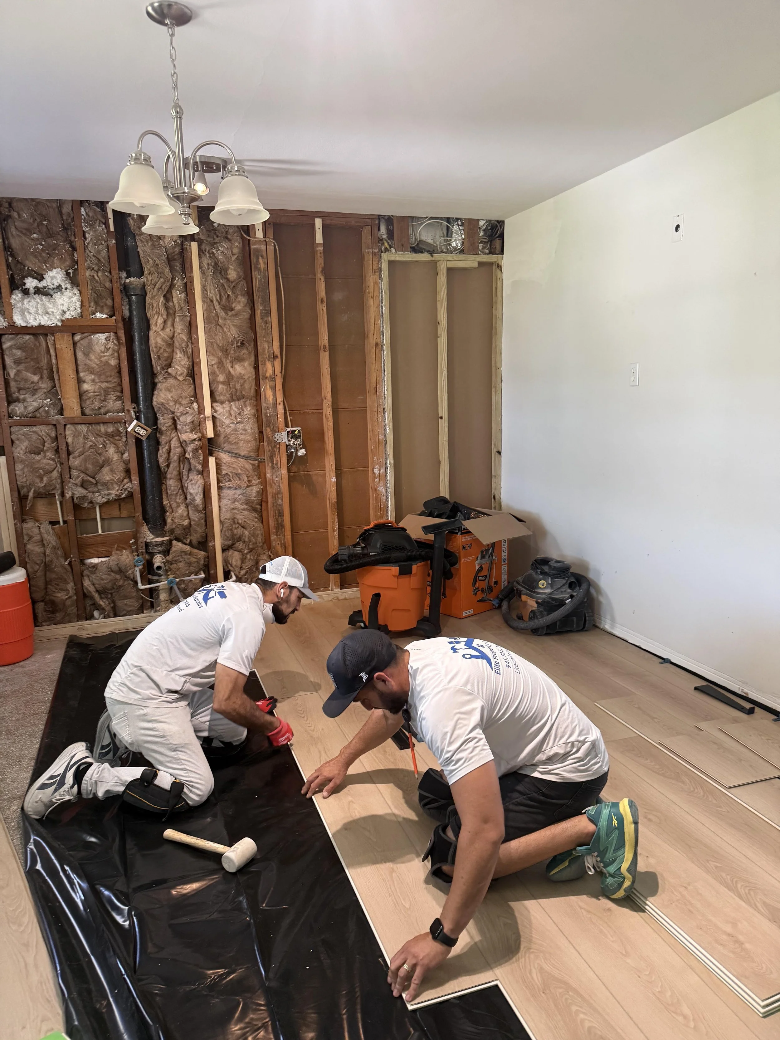 Two men are kneeling on the floor installing a piece of wood or flooring. One man is wearing a white t-shirt, white cap, and gloves, while the other is wearing a gray cap. Various tools and materials are scattered around them. The room appears to be under renovation, with exposed wall studs and insulation visible.