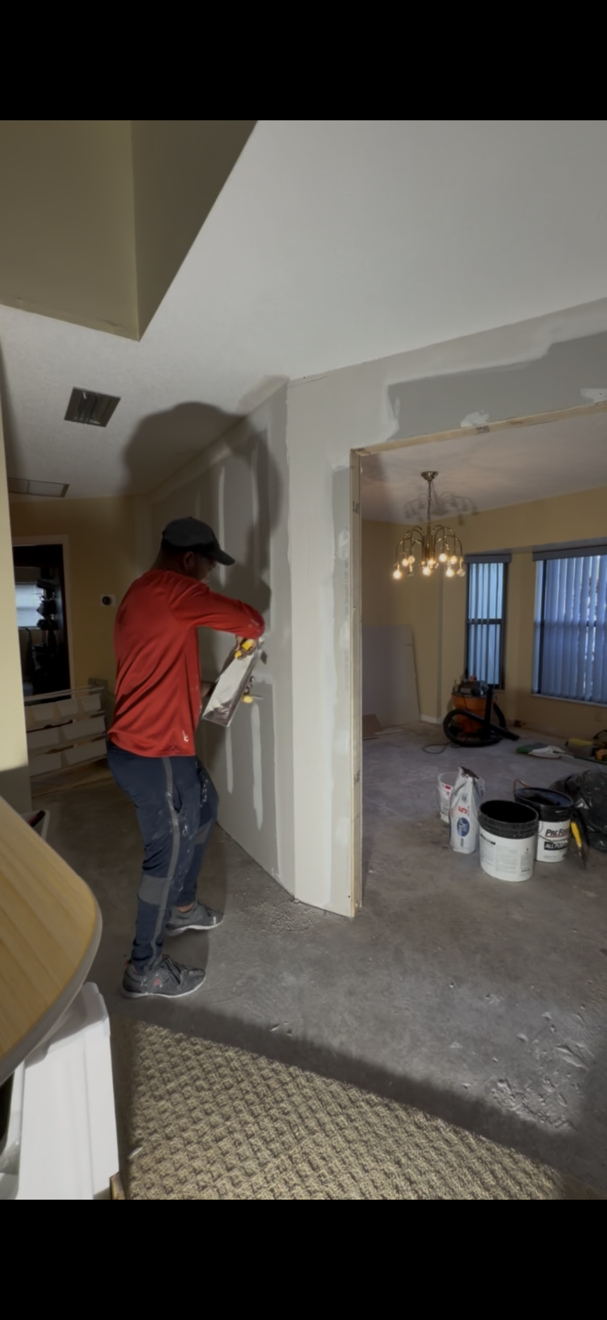 A person in a red hoodie is applying drywall mud to a doorway in a room under construction, with construction tools, paint buckets, and a vacuum cleaner on the floor.