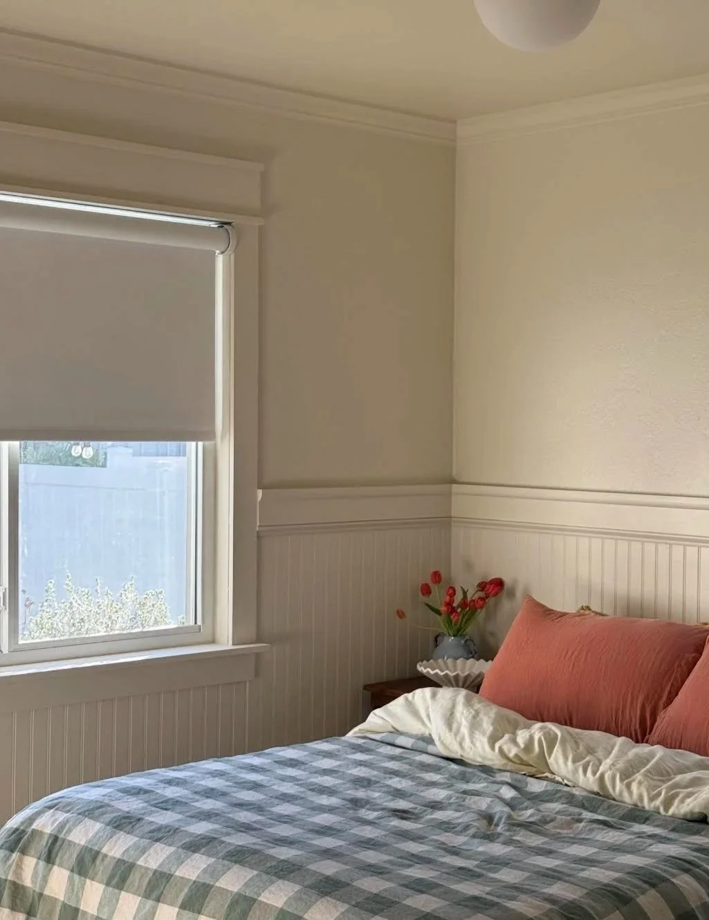 A cozy bedroom with a bed that has pink pillows and a checkered bedspread. There is a window with a closed white shade, and a small side table with a vase of red flowers.