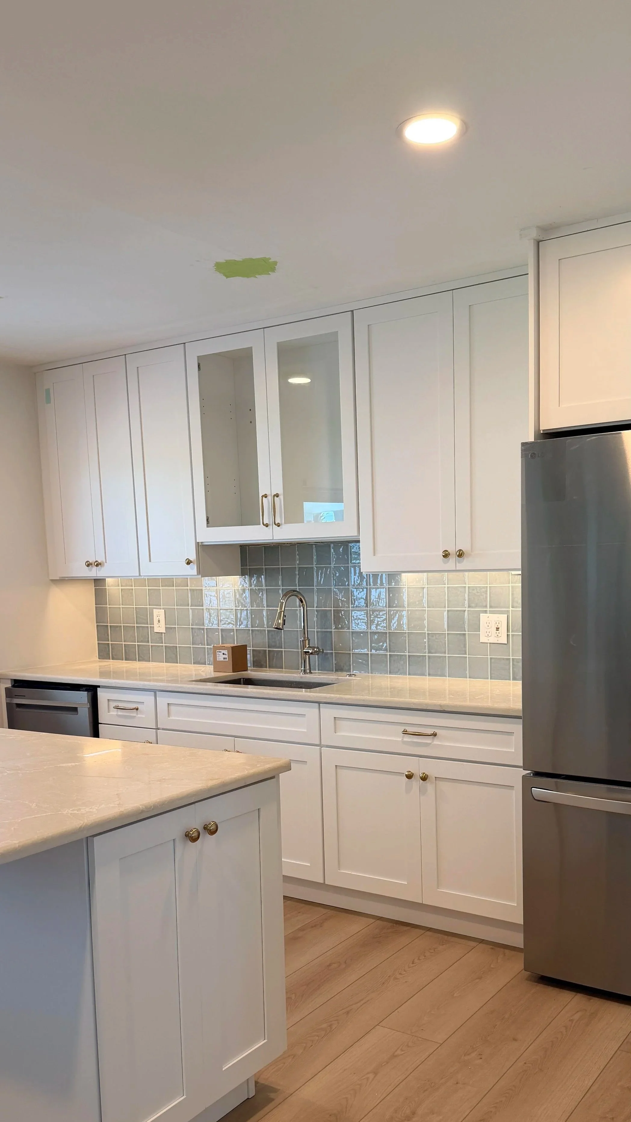 Modern kitchen with white cabinetry, light wood flooring, gray tiled backsplash, stainless steel refrigerator, and under-cabinet lighting.