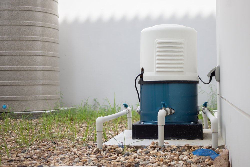 Outside air conditioning unit with pipes connected, surrounded by gravel and grass, located near a wall and a water tank.