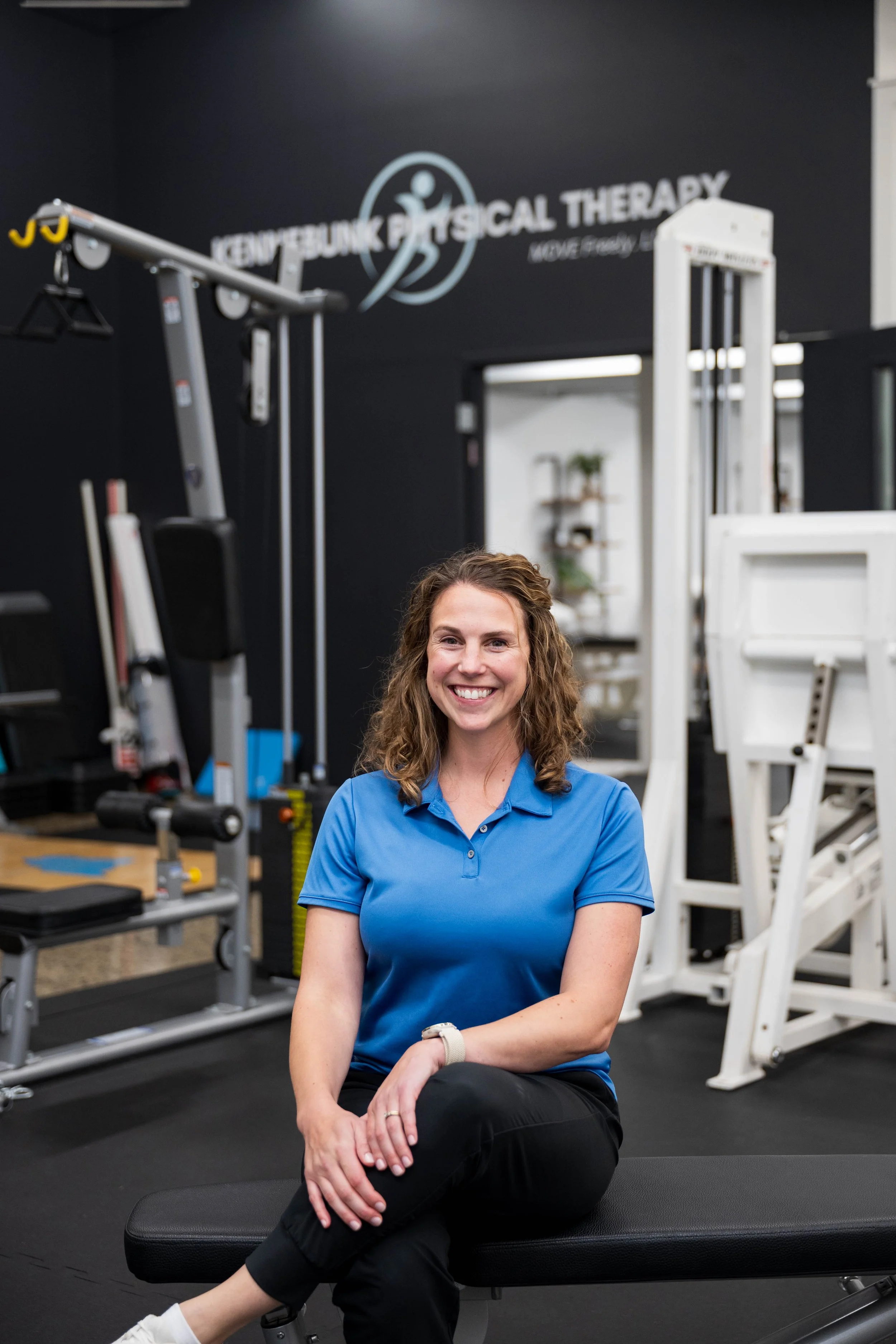 Smiling physical therapist in a blue polo seated on a treatment table in a physical therapy gym