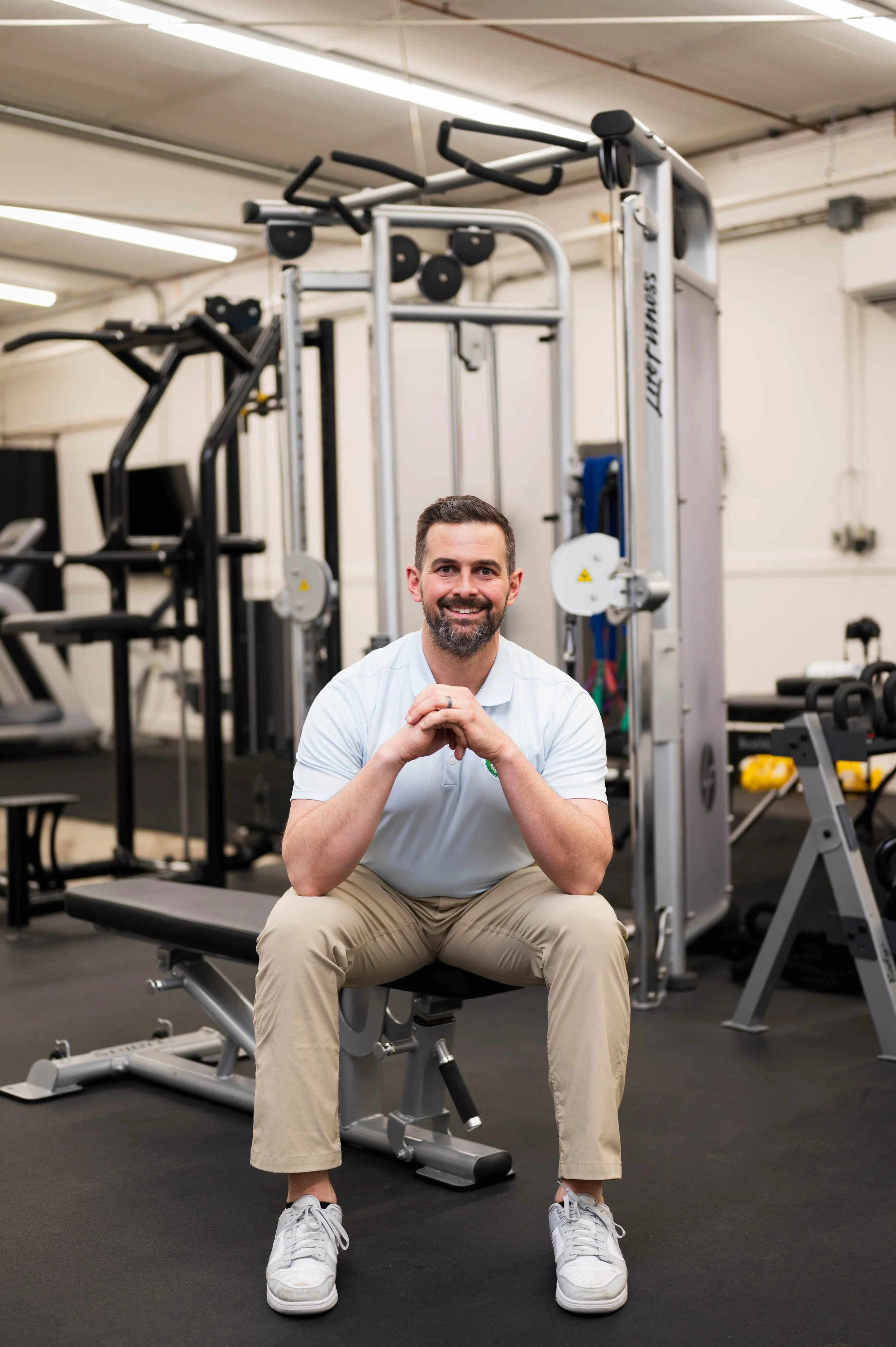 Smiling man seated on a weight bench in a gym surrounded by strength-training equipment.