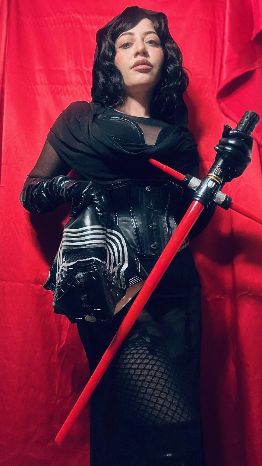 A woman with black wavy hair dressed in a dark outfit, holding a red staff, standing against a red curtain.