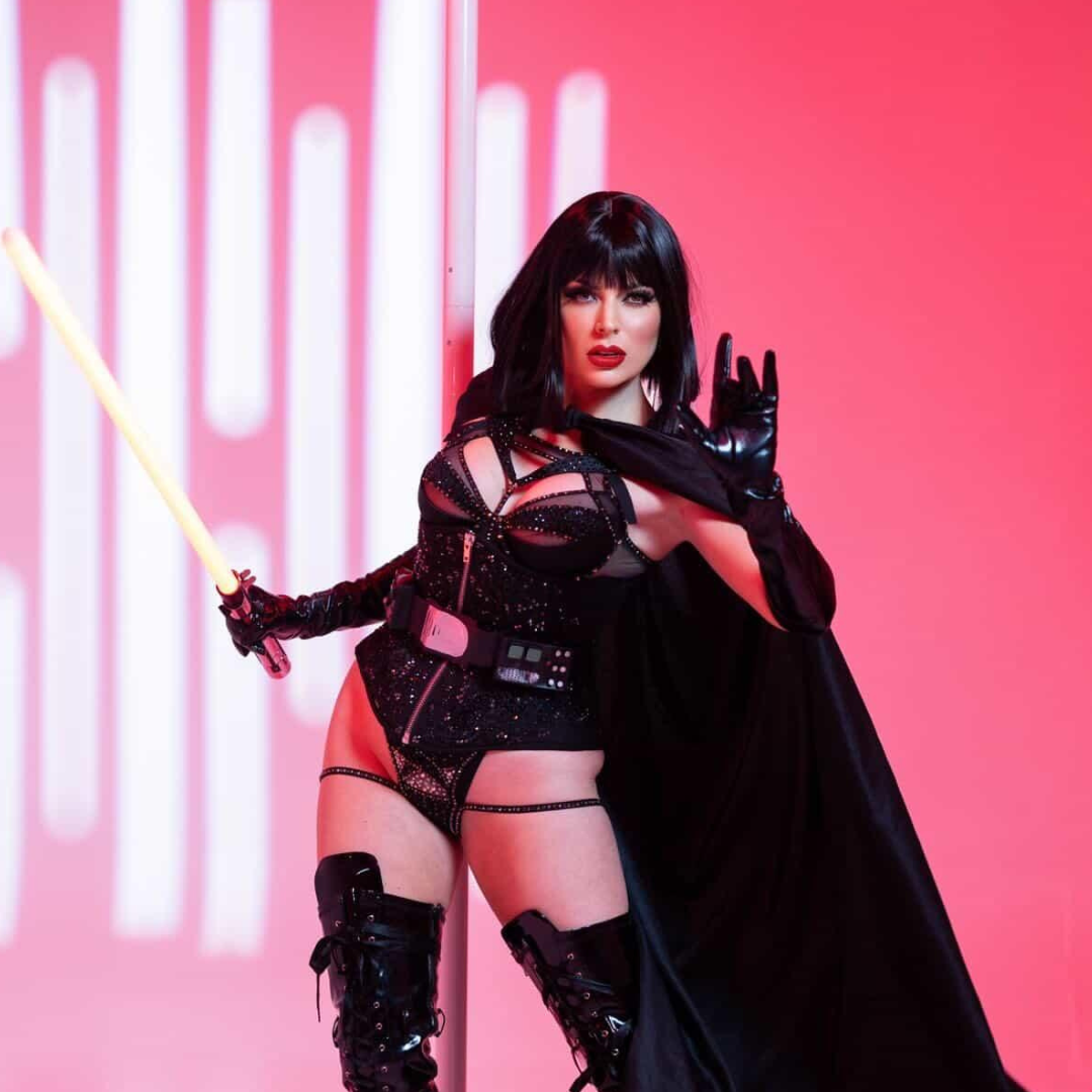 Woman dressed as a sexy Star Wars character in a black costume with shiny elements, holding a lightsaber, standing in front of a pink background.