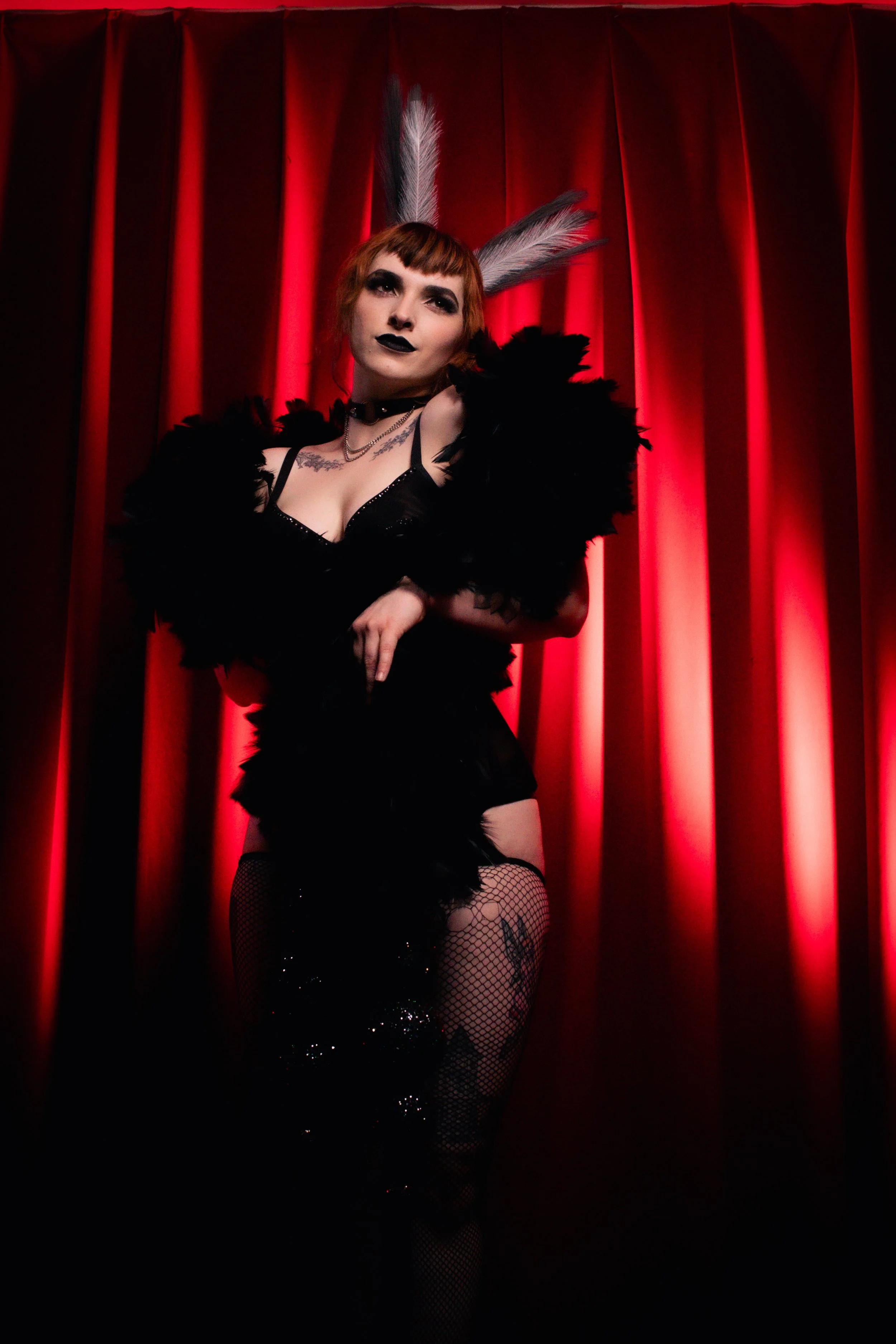 A woman with short red hair, dark makeup, and feathers in her hair, standing in front of red curtains, wearing Gothic-style black clothing, fishnet stockings, and jewelry.