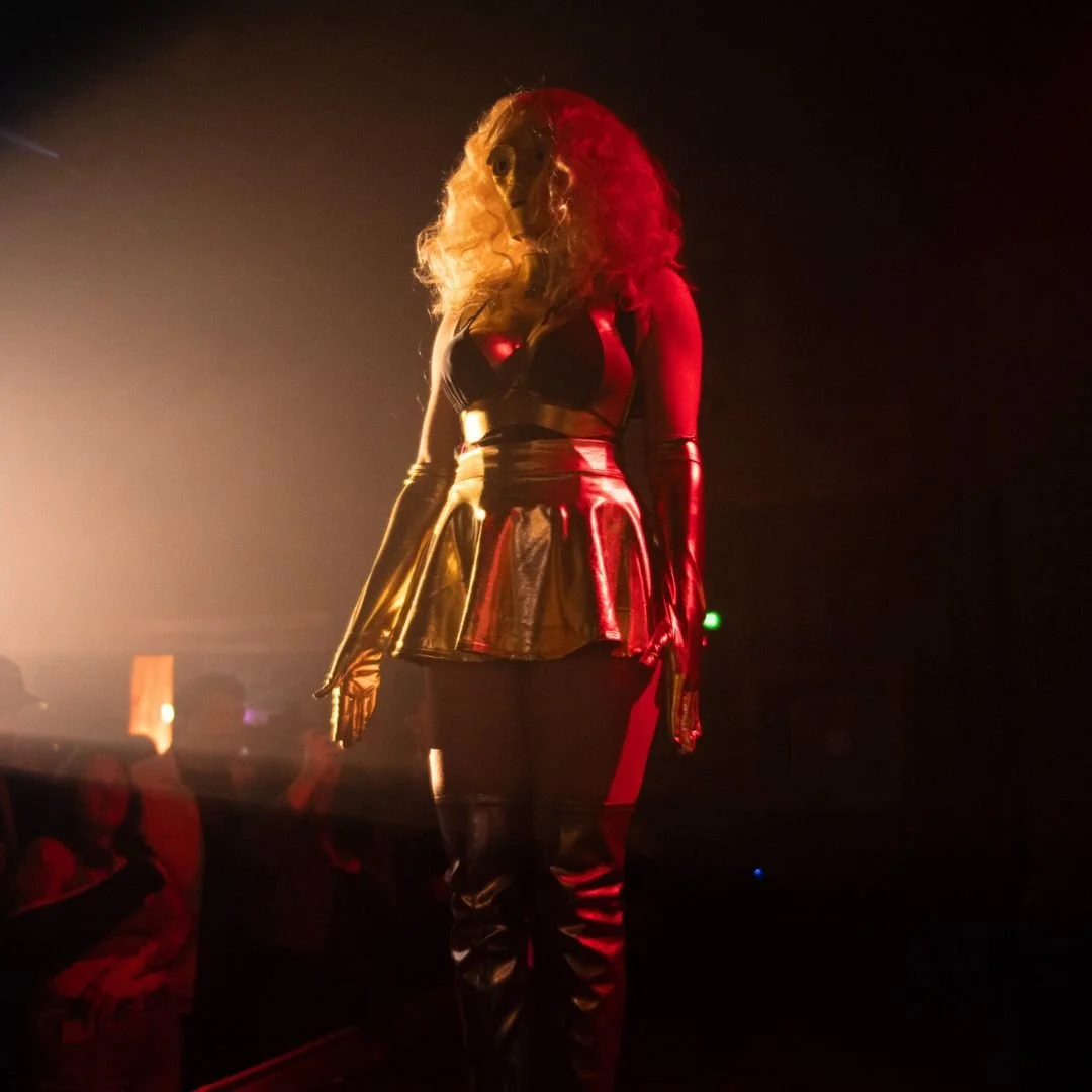 A person wearing a black mask, a shiny black top, a gold skirt, and long gloves, standing in a dark, smoky environment with red and yellow lighting.