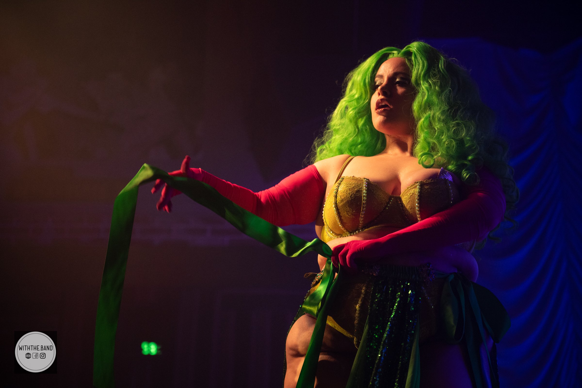 Performer with bright green curly hair and gold top holding a green ribbon on stage with dark background and colorful lighting.