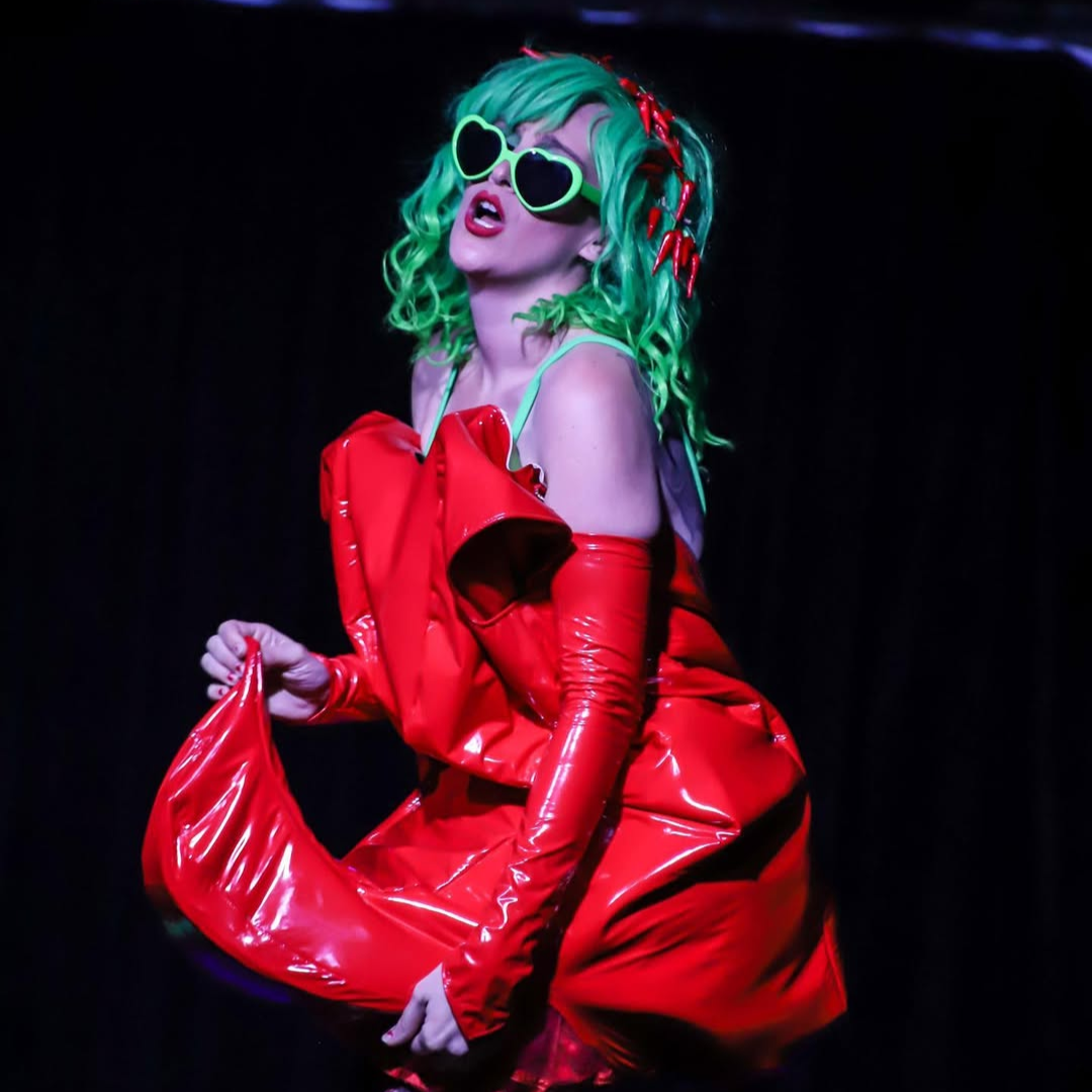 Performer with neon green hair, sunglasses, and red shiny outfit, singing on stage.