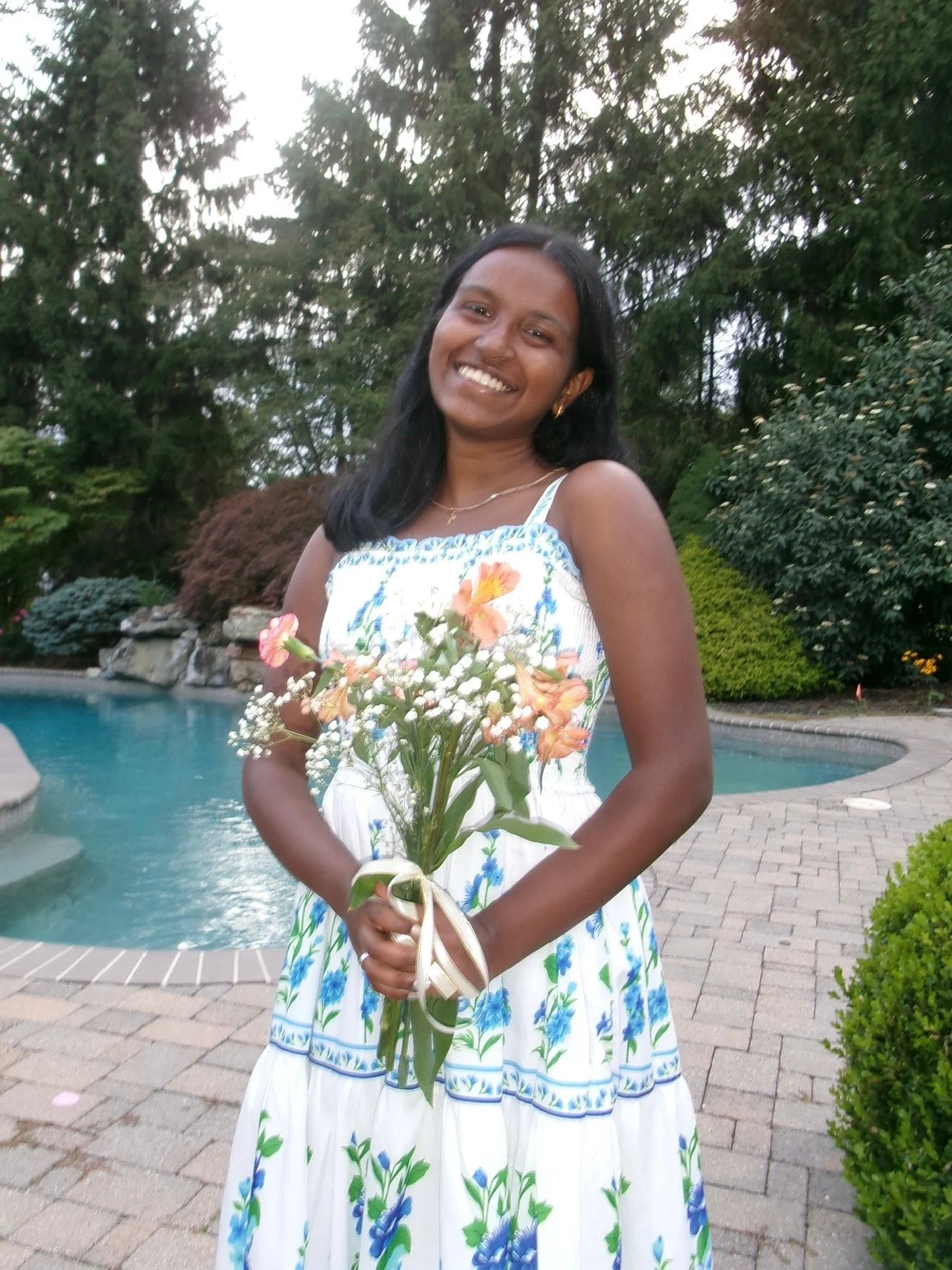 Smiling person with dark hair in a blue shirt, outdoors with green foliage in the background.