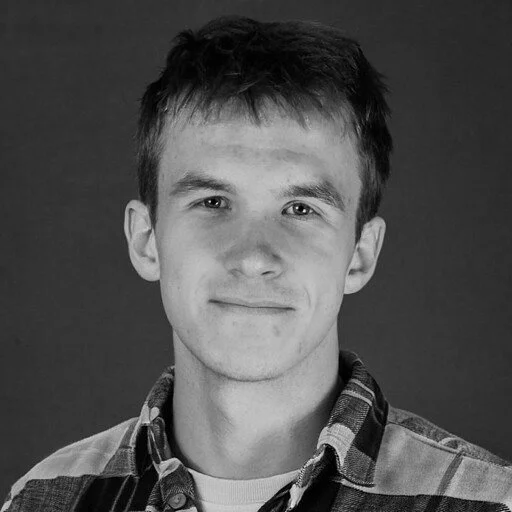 Black and white portrait of a young man with short hair, wearing a checkered shirt, smiling slightly against a plain dark background.