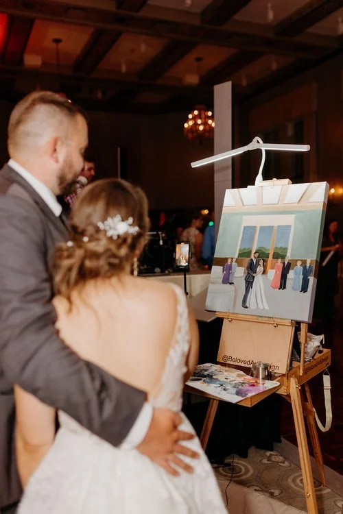 A couple in wedding attire is standing in front of an easel holding a painting of their wedding. The painting depicts the couple dancing with guests in the background.