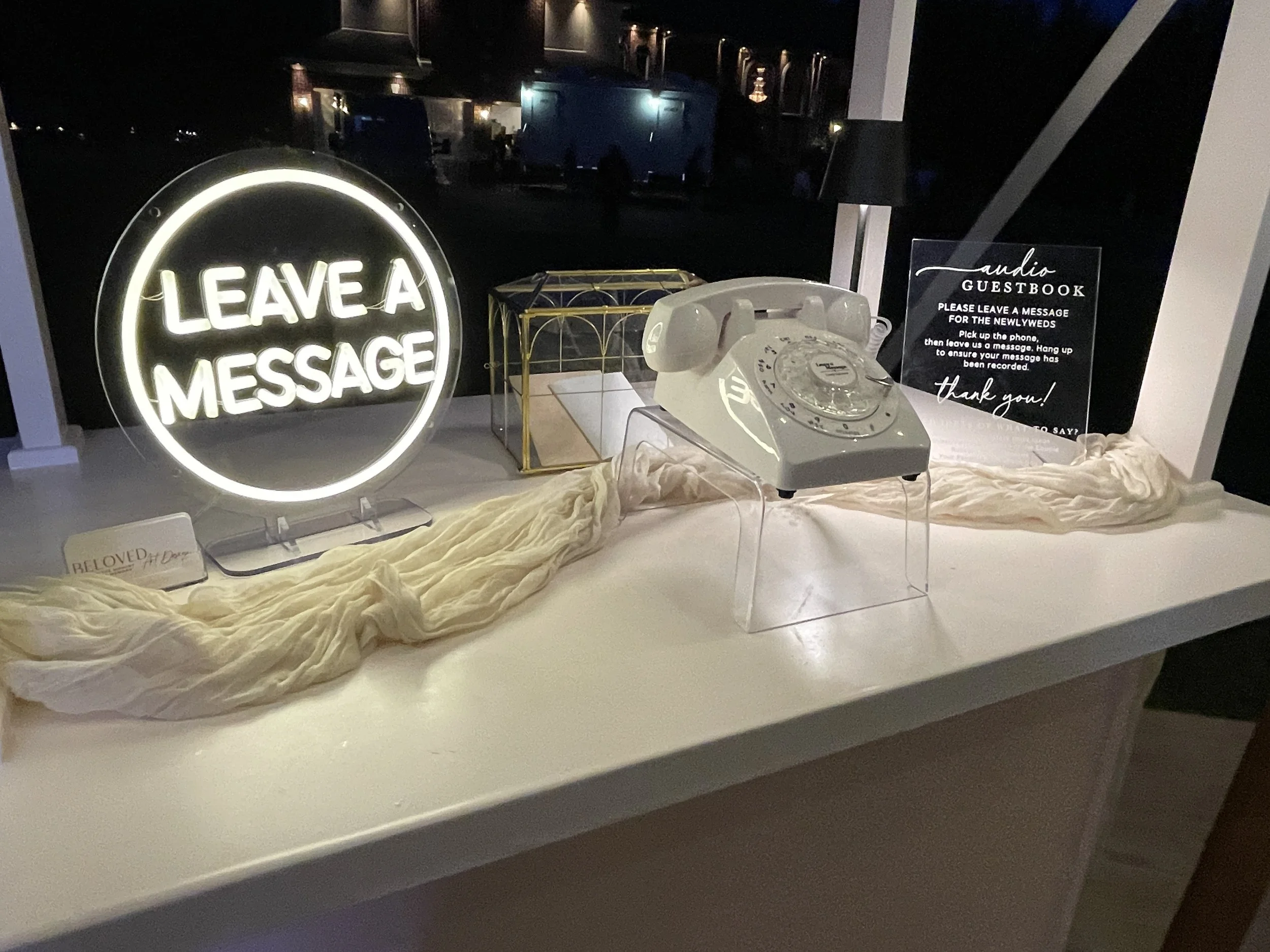 A neon sign that says 'Leave a Message' on a white surface, a vintage rotary telephone, a guestbook sign, and a decorative gold wire basket on a table with a cream-colored cloth. The background is dark with faint reflections of lights.