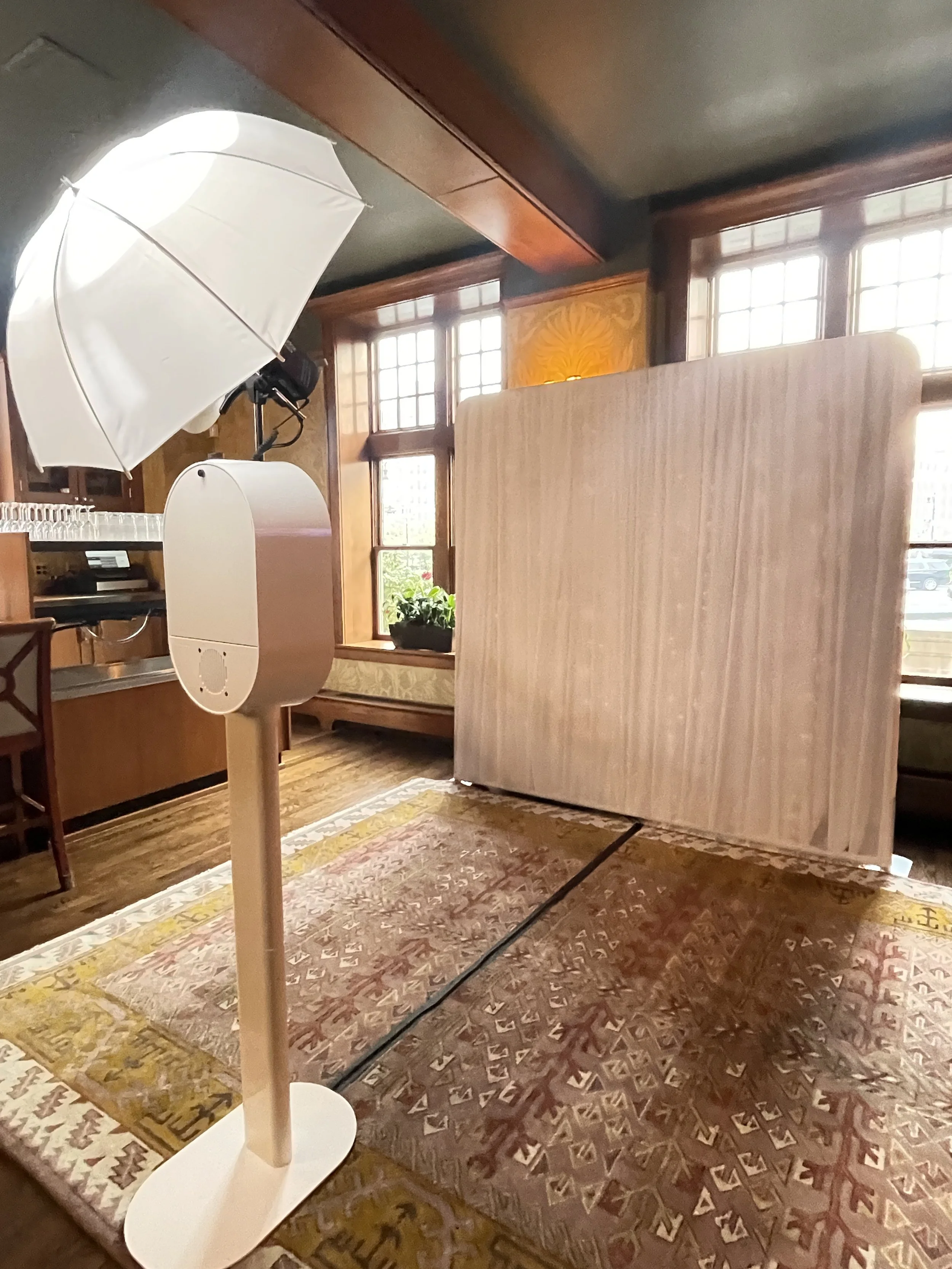 Indoor photo booth setup with umbrella light and wooden backdrop in a warmly lit room with large windows and patterned rugs.