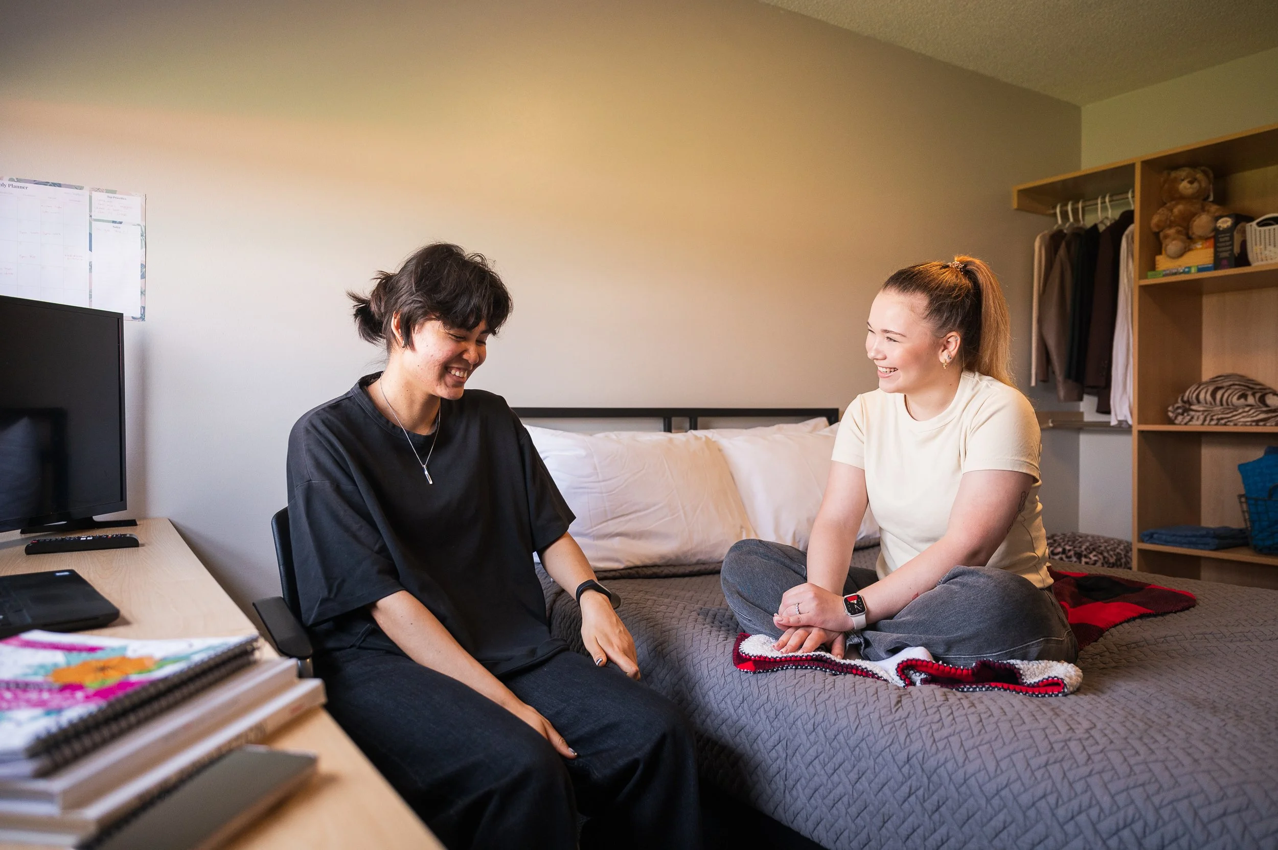 Two women sitting on a bed in a bedroom, smiling and laughing together.