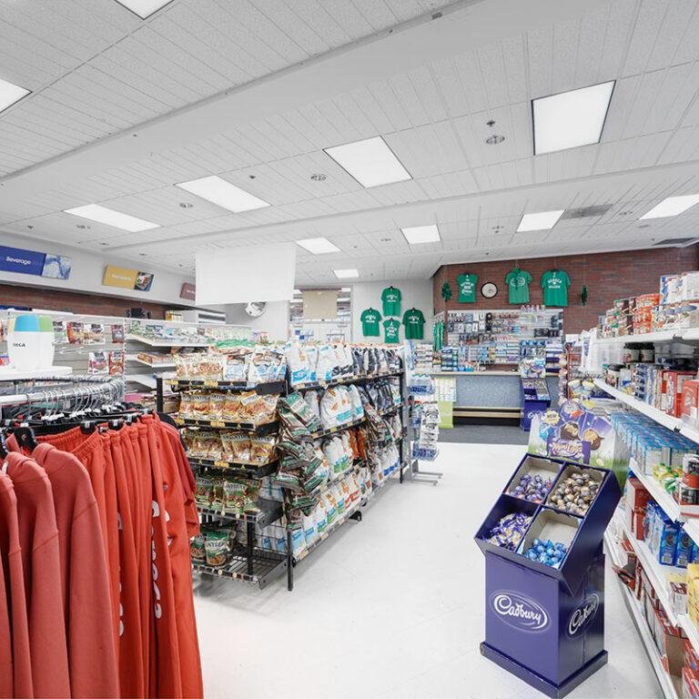 Inside a retail store with shelves stocked with snacks, candy, and merchandise, including red sweatshirts hanging on the left and a display of assorted chocolates in the center, with a checkout counter in the background.