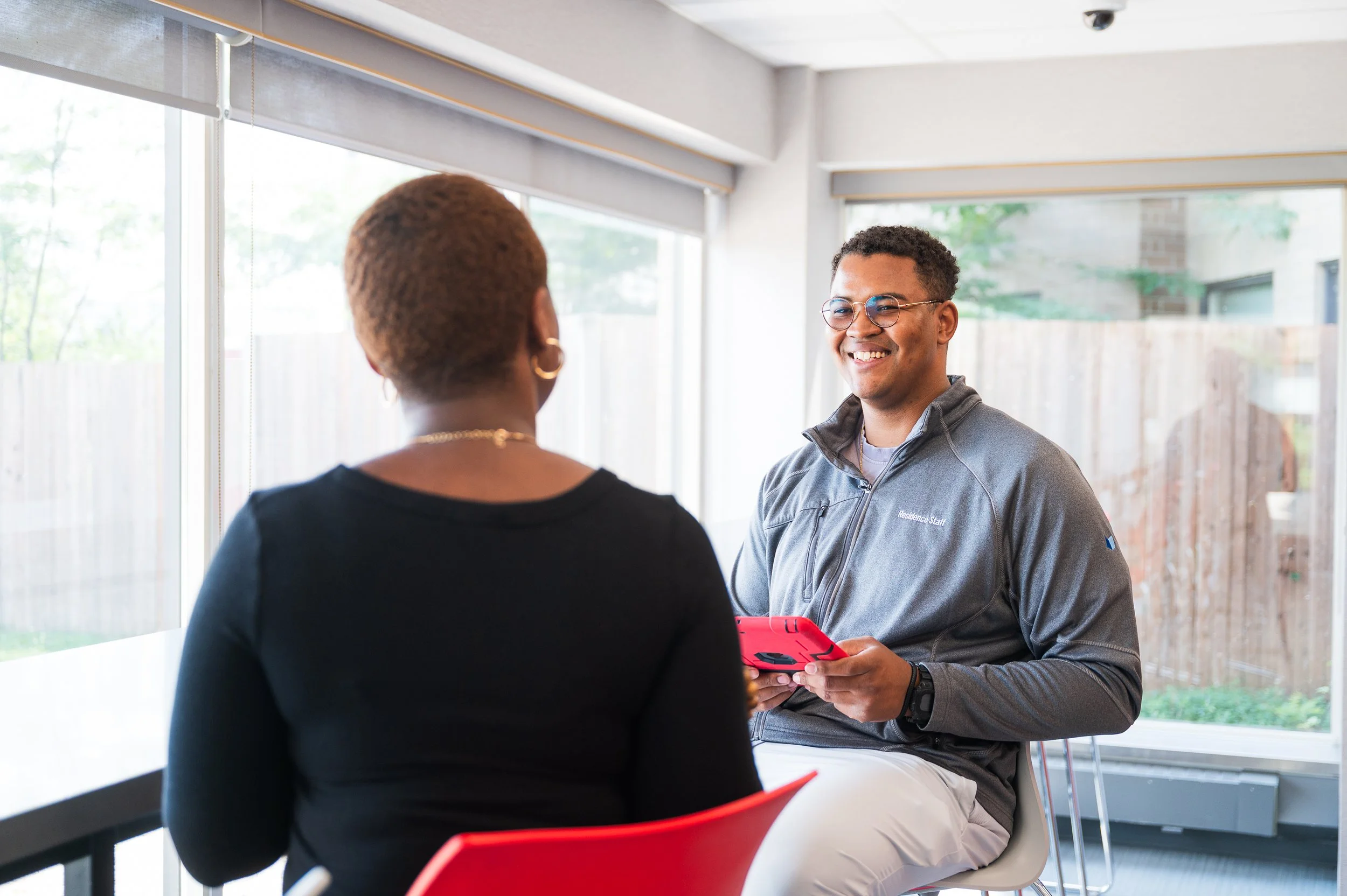 A young man with glasses and a gray jacket smiling while holding a red smartphone, sitting in front of a woman with short hair in a black top.