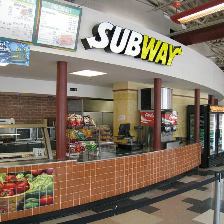 SUBWAY fast food restaurant counter with menu boards, snack shelves, soda dispenser, and refrigerated drinks in the background.