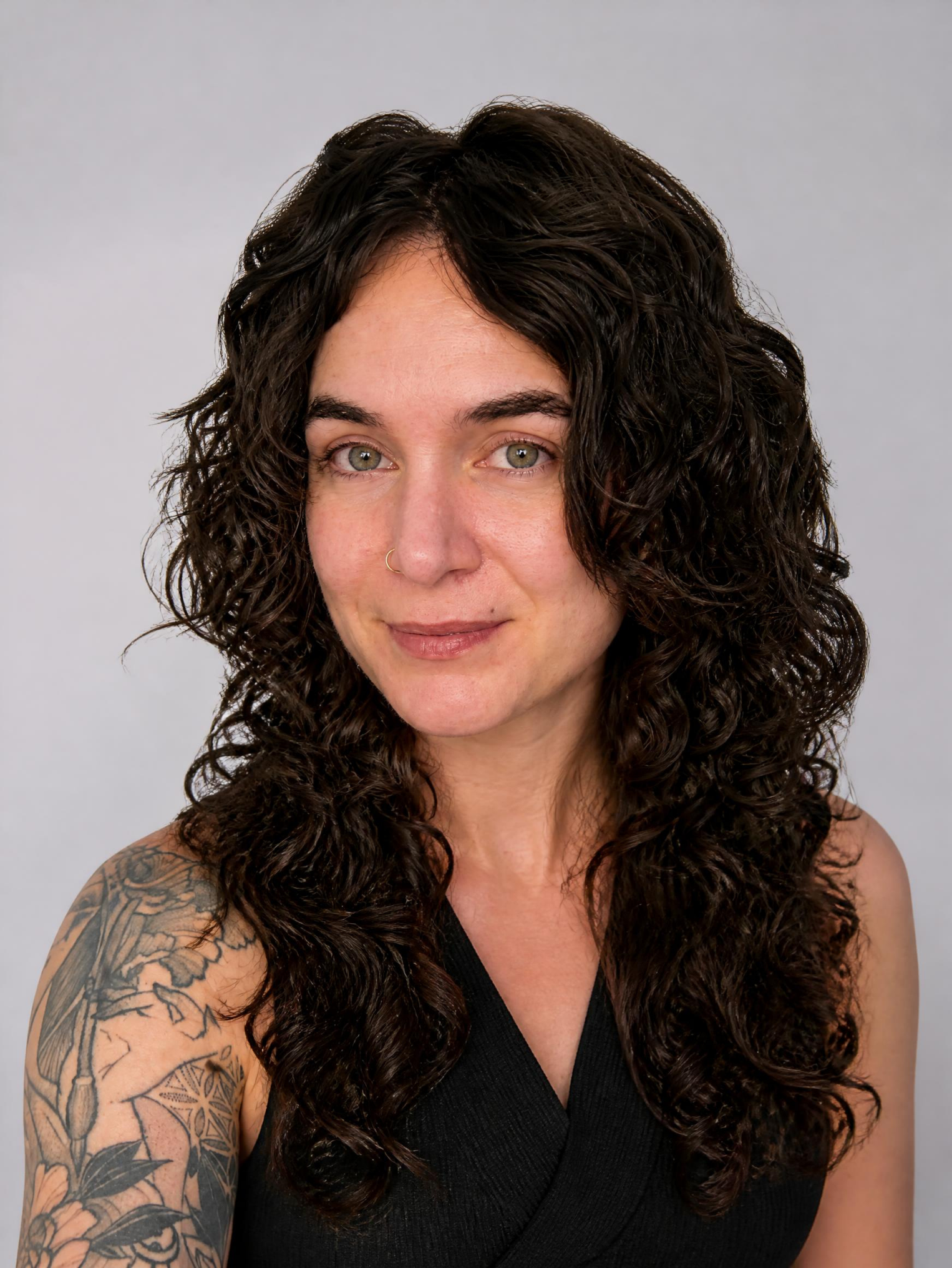 Close-up portrait of a woman with curly dark hair, tattoos on her left arm, and a nose ring, wearing a black top, against a plain gray background.