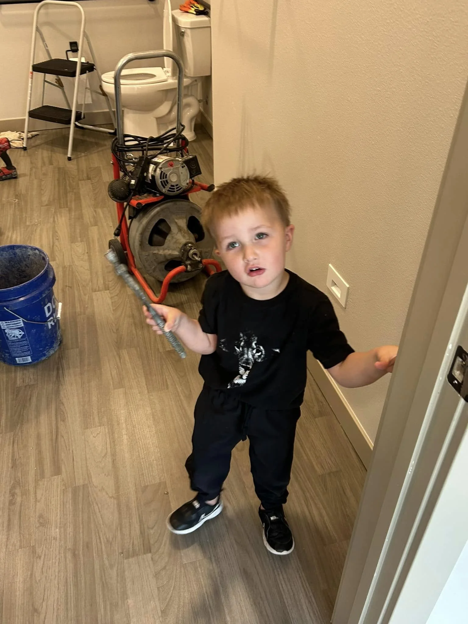 Young child holding a tool in a room with plumbing equipment and a toilet, wearing black clothes and sneakers.